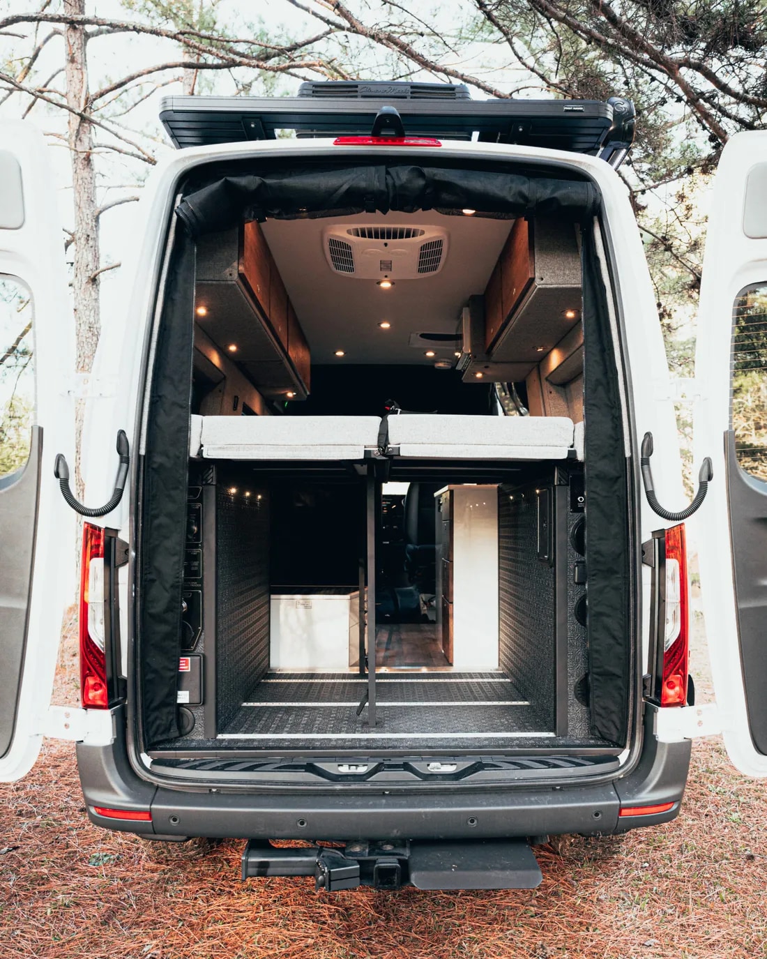 An rv rental camper van's open rear reveals a smart interior with raised bed, lower storage, and ceiling AC unit.. Mercedes-Benz Sprinter 2022