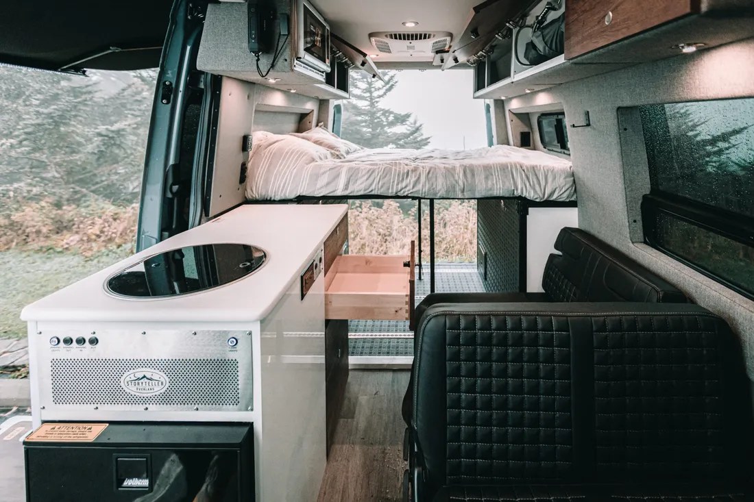 This rv rental offers a compact interior with a raised bed, kitchen, seating, & nature view through open rear doors.. Mercedes-Benz Sprinter 2022