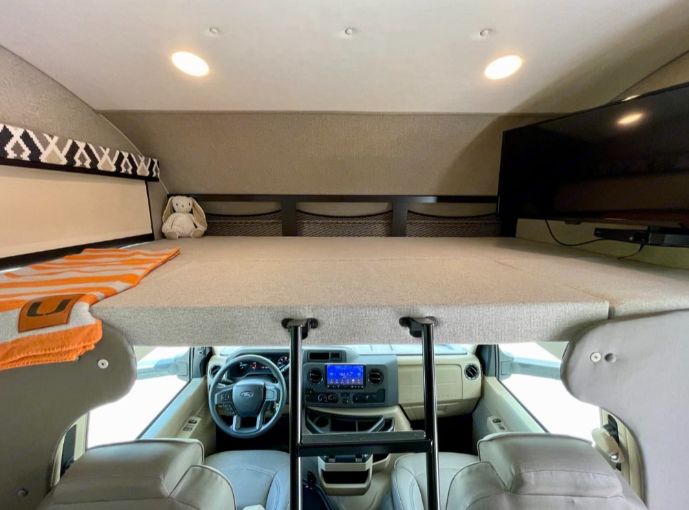 Interior of an RV rental showing the comfortable over-cab bunk bed with a ladder, overlooking the Ford cab and dashboard.. Jayco Greyhawk 2021