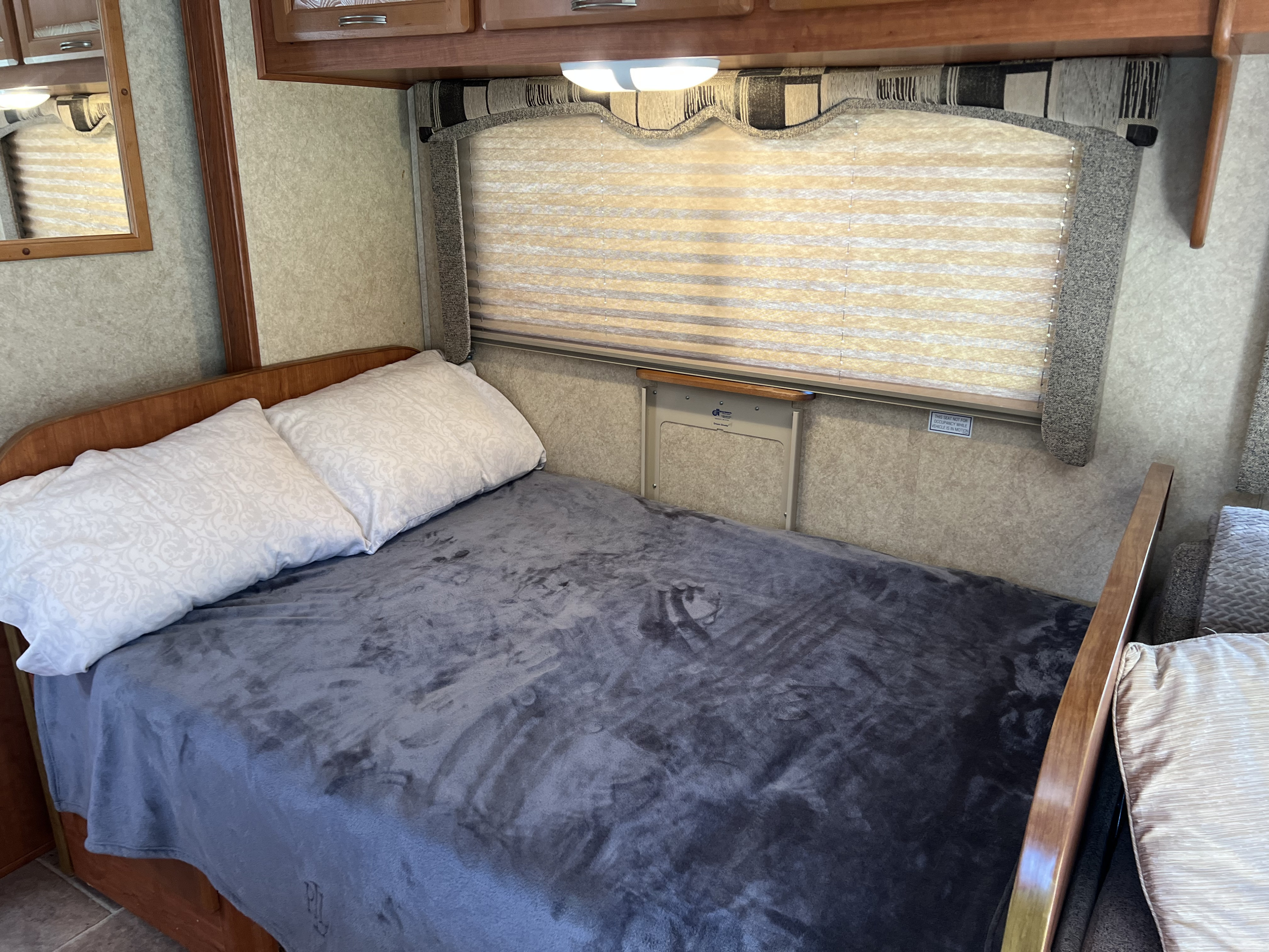 RV rental interior: a neatly made bed with white pillows and a grey blanket, under a window with blinds and wood cabinets.. Forest River Sunseeker 2010