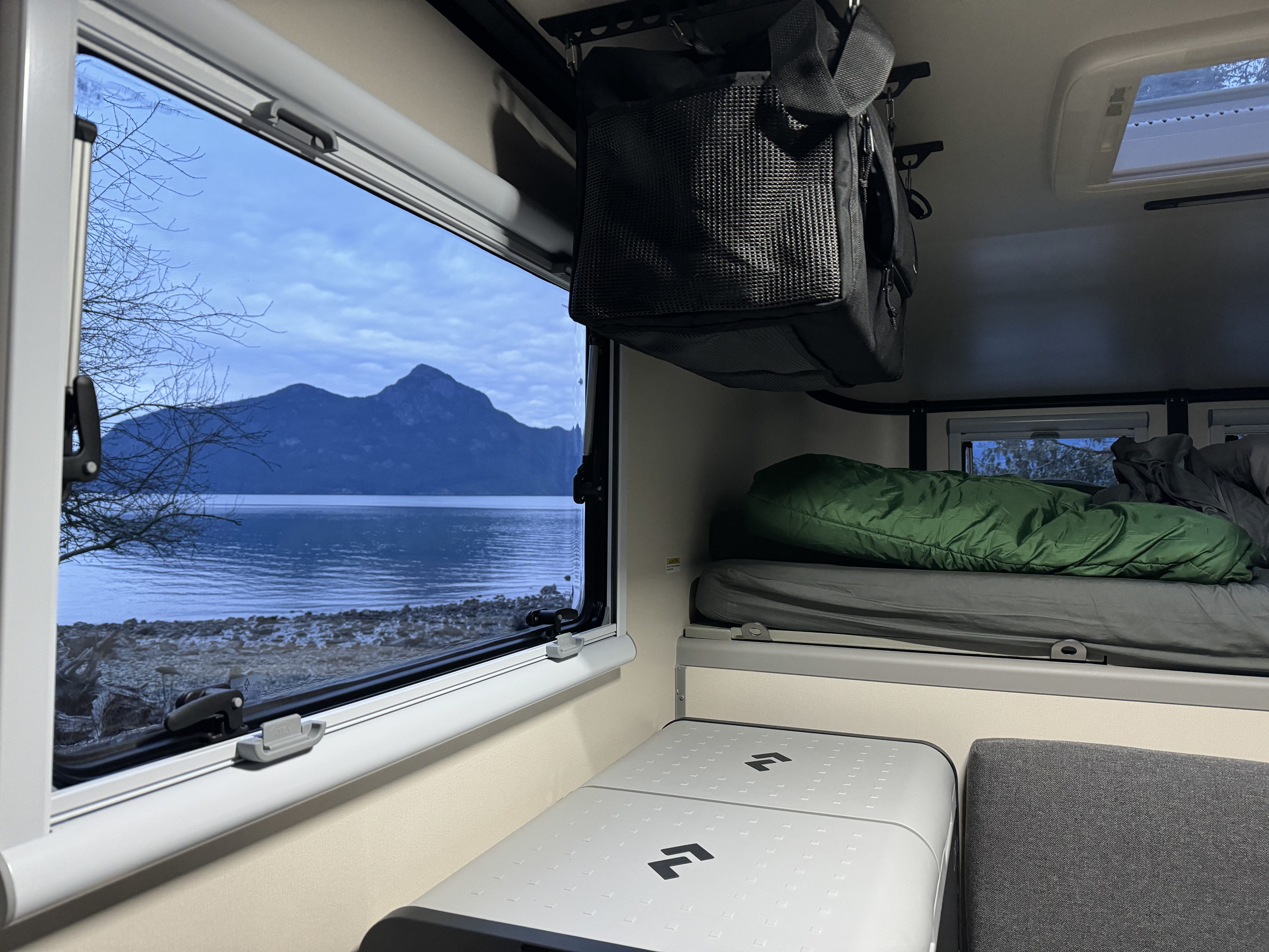 RV rental interior: A bed, portable fridge, and hanging storage face a window overlooking a tranquil lake, mountain, and bare trees.. Dodge Dodge 1 Ton Pu 2018
