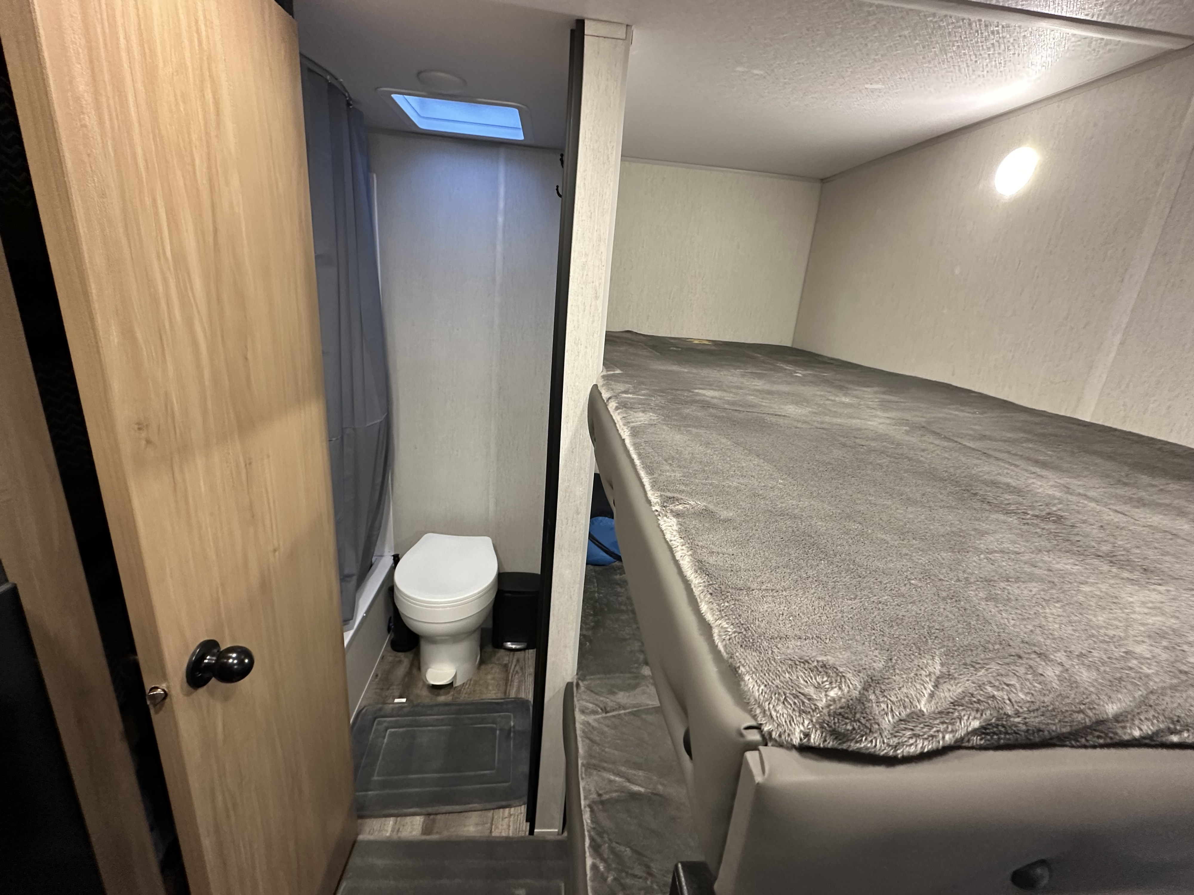 Compact RV rental interior: small bathroom with toilet and shower, adjacent to a bunk bed with a fuzzy gray blanket.. Forest River Cherokee Wolf Den 16 EV 2024