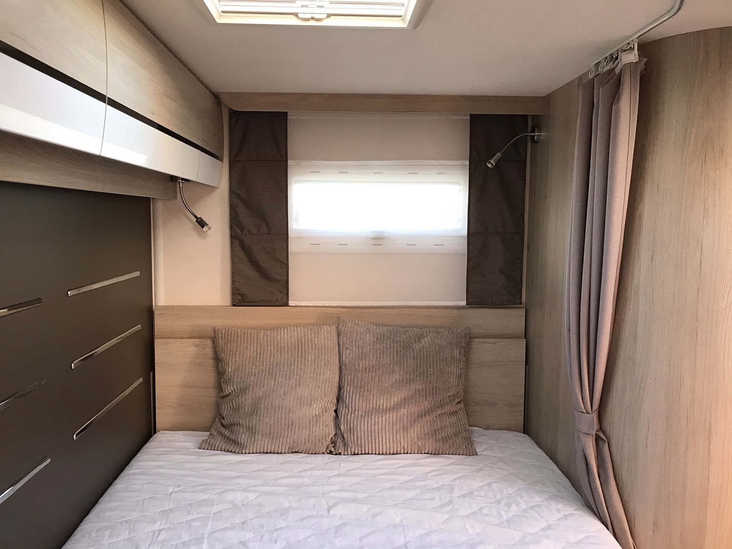 An RV rental bedroom features a comfy bed with pillows, wooden headboard, window blinds, and storage cabinetry on the left.. Chausson 514 2019