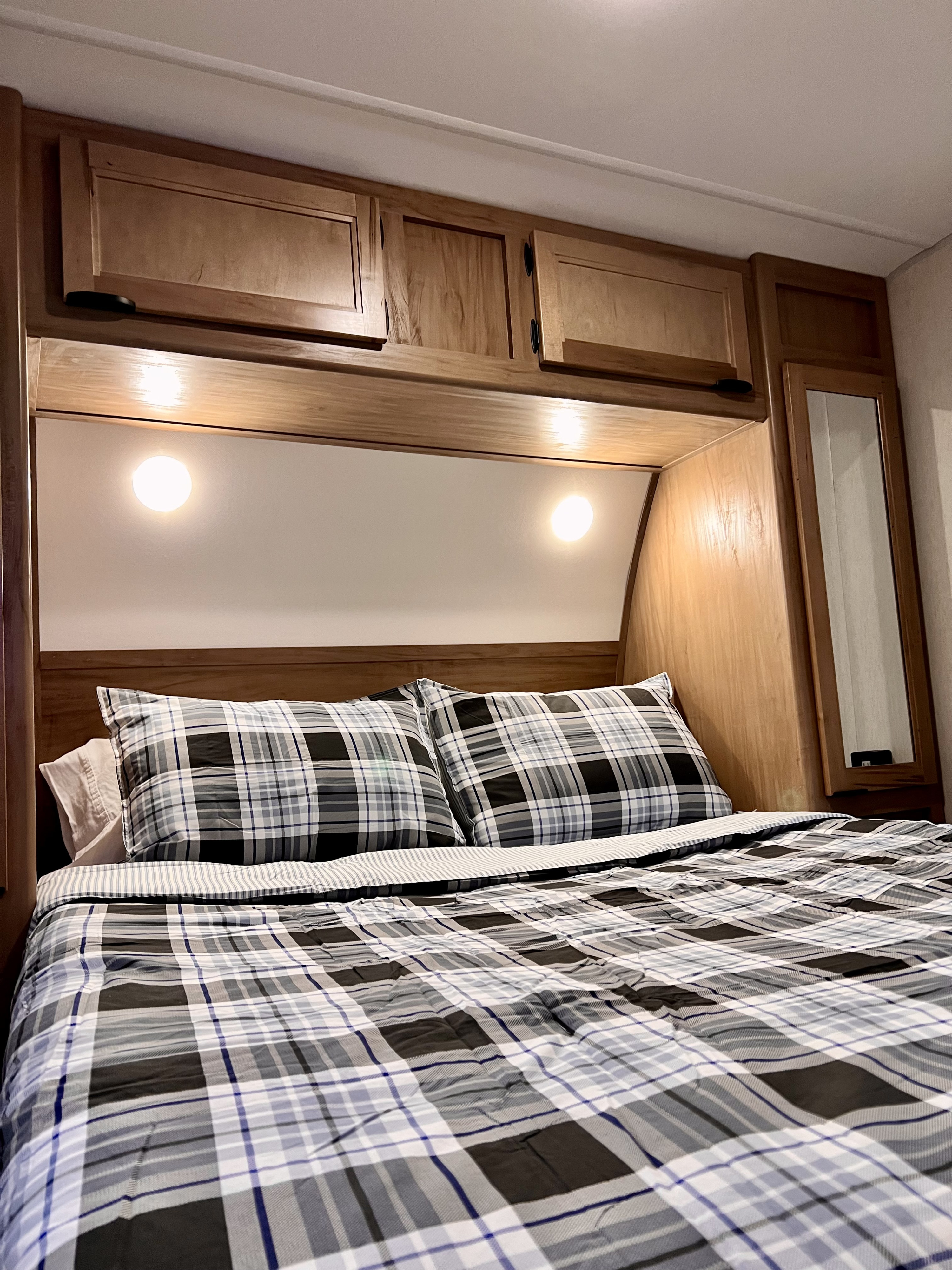 Experience comfort in this RV rental bedroom, featuring plaid bedding, convenient overhead storage, and warm lighting.. Forest River Cherokee Grey Wolf 2024