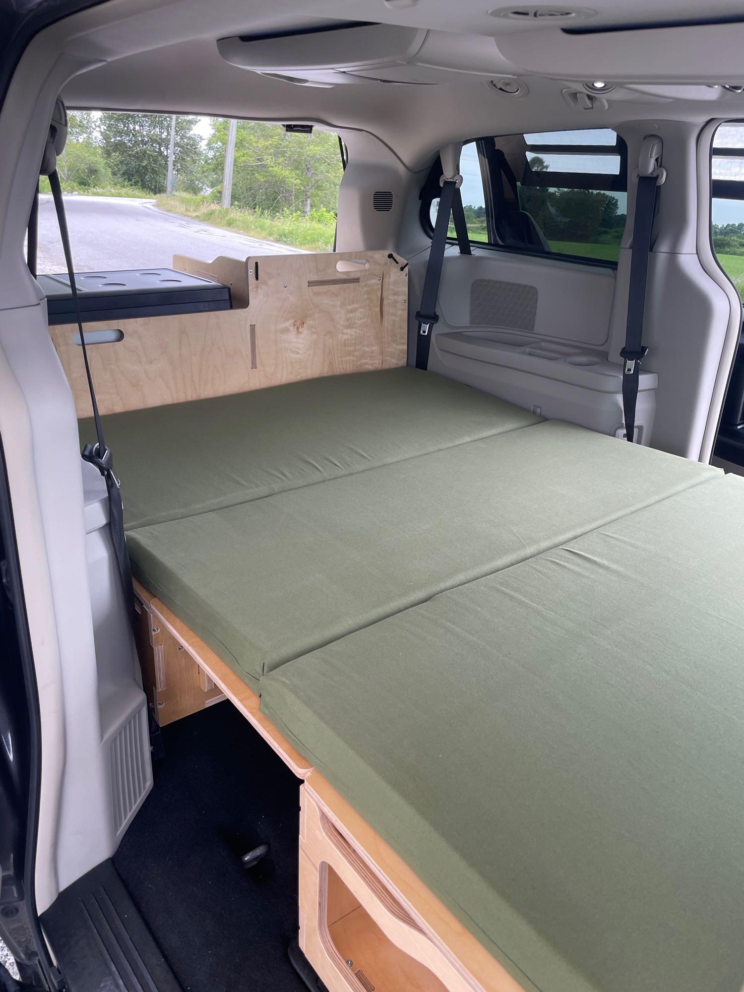 This RV rental features a converted minivan interior with a wooden bed platform, green mattress, and cooler, ready for camping.. Dodge Grand Caravan 2015