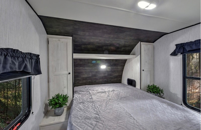 Cozy RV rental bedroom with a quilted bed, dark wood headboard, side cabinets, and outside forest views from windows.. Heartland RVs Trail Runner 2022