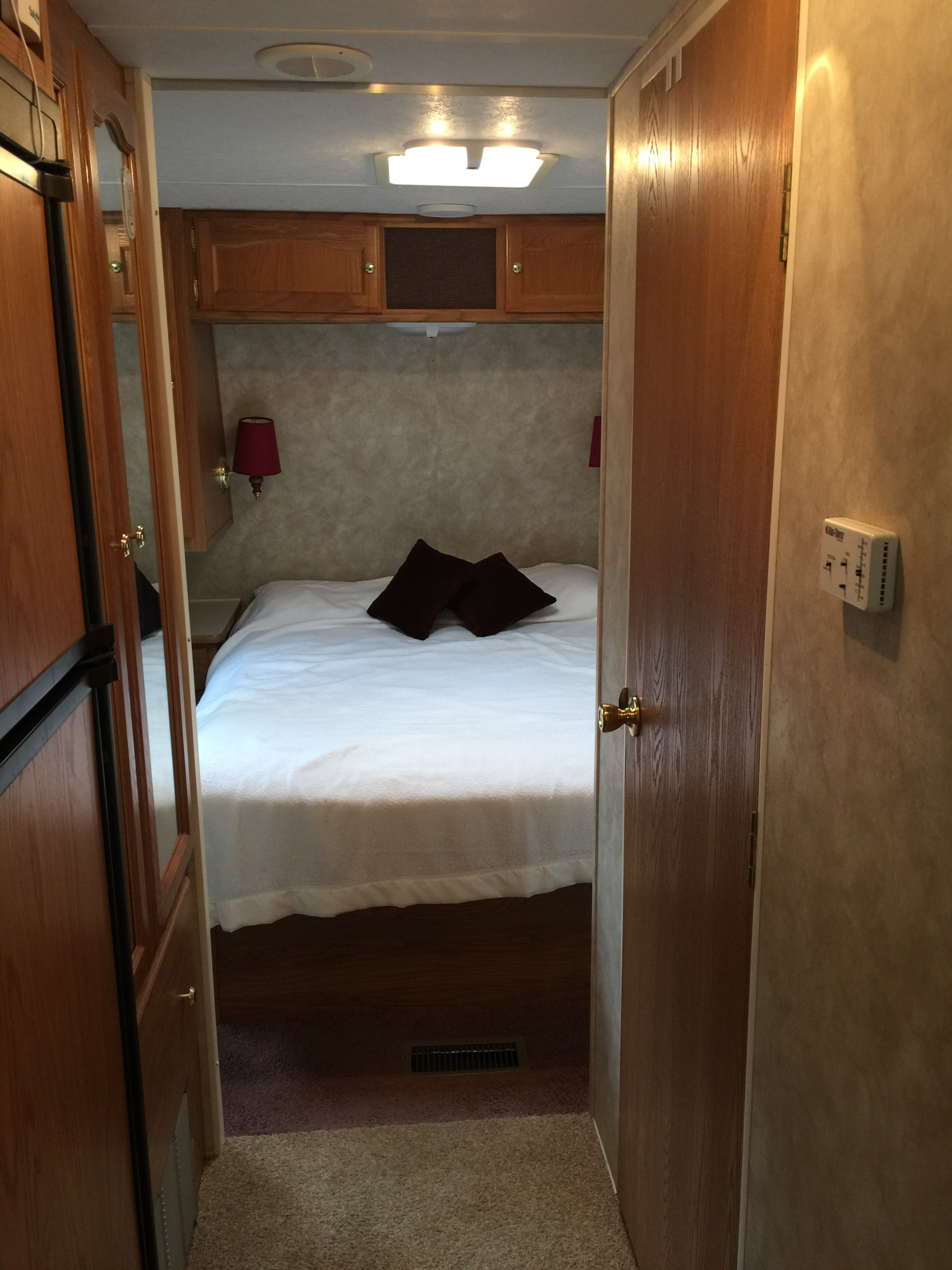 View into an RV rental bedroom: white bed with dark pillows, wall lamps, and wood cabinetry, seen through an open doorway.. Keystone RV Springdale 2005