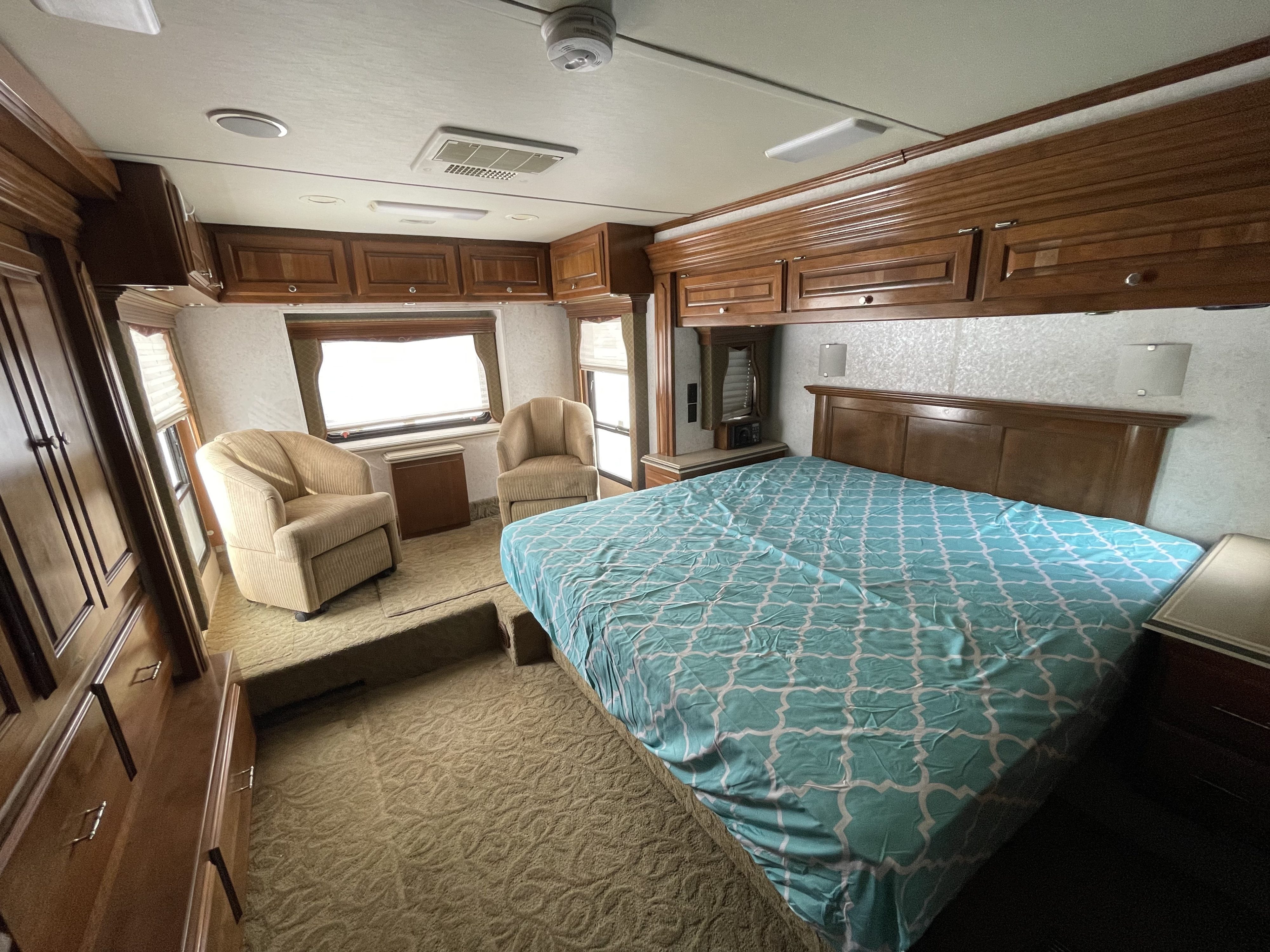 Spacious RV rental bedroom featuring a teal patterned bed, ample wood cabinets, and two cozy armchairs for a comfortable motorhome stay.. Monaco Camelot 2007
