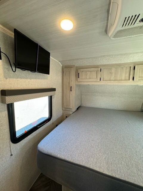 Inside a clean RV rental bedroom, featuring a large bed, wall-mounted TV, window, and wood-look overhead cabinets.. East to West Delta Terra 2022