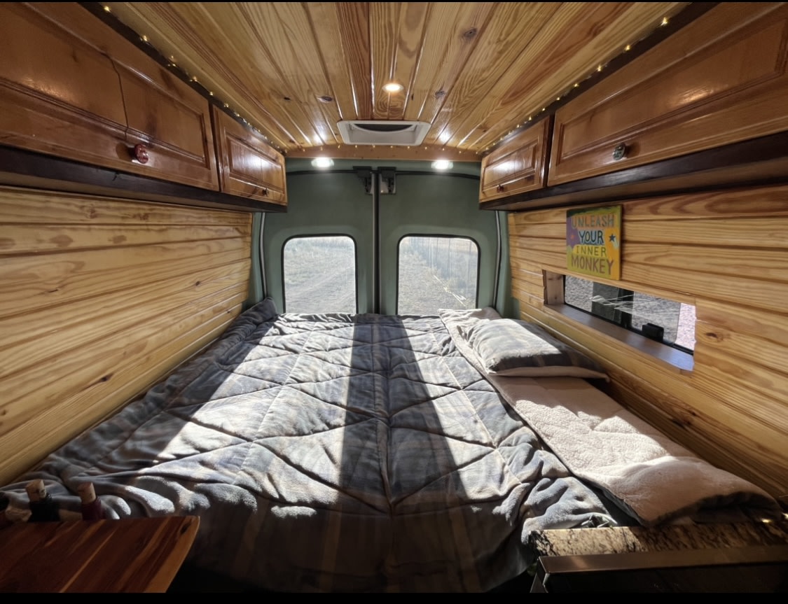 A comfortable **RV rental** interior showcasing a large bed, warm wood paneling, and natural light from rear windows.. Ford Transit 350 High Roof EXT 2020