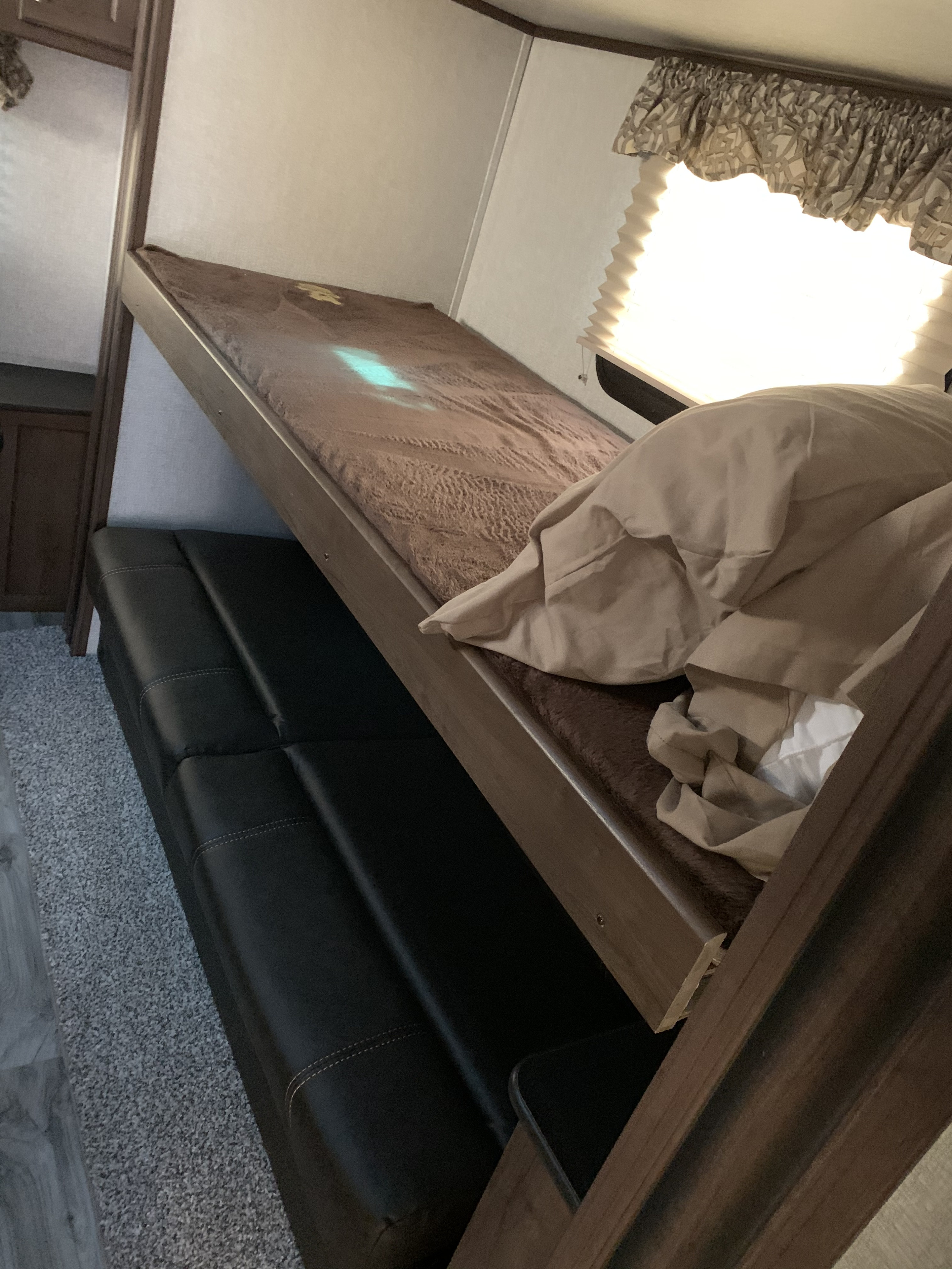 Inside an RV rental: a top bunk bed with brown mattress and a black sofa below. A window with shades is on the right.. Keystone RV Cougar 2019