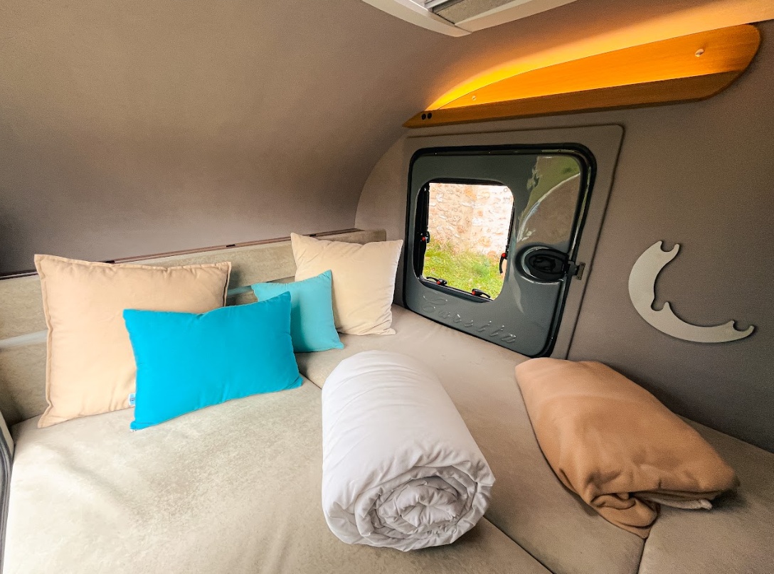 Cozy RV rental sleeping area featuring a bed, beige and blue pillows, and blankets. A small window offers an outdoor view.. Caretta 1500 2024