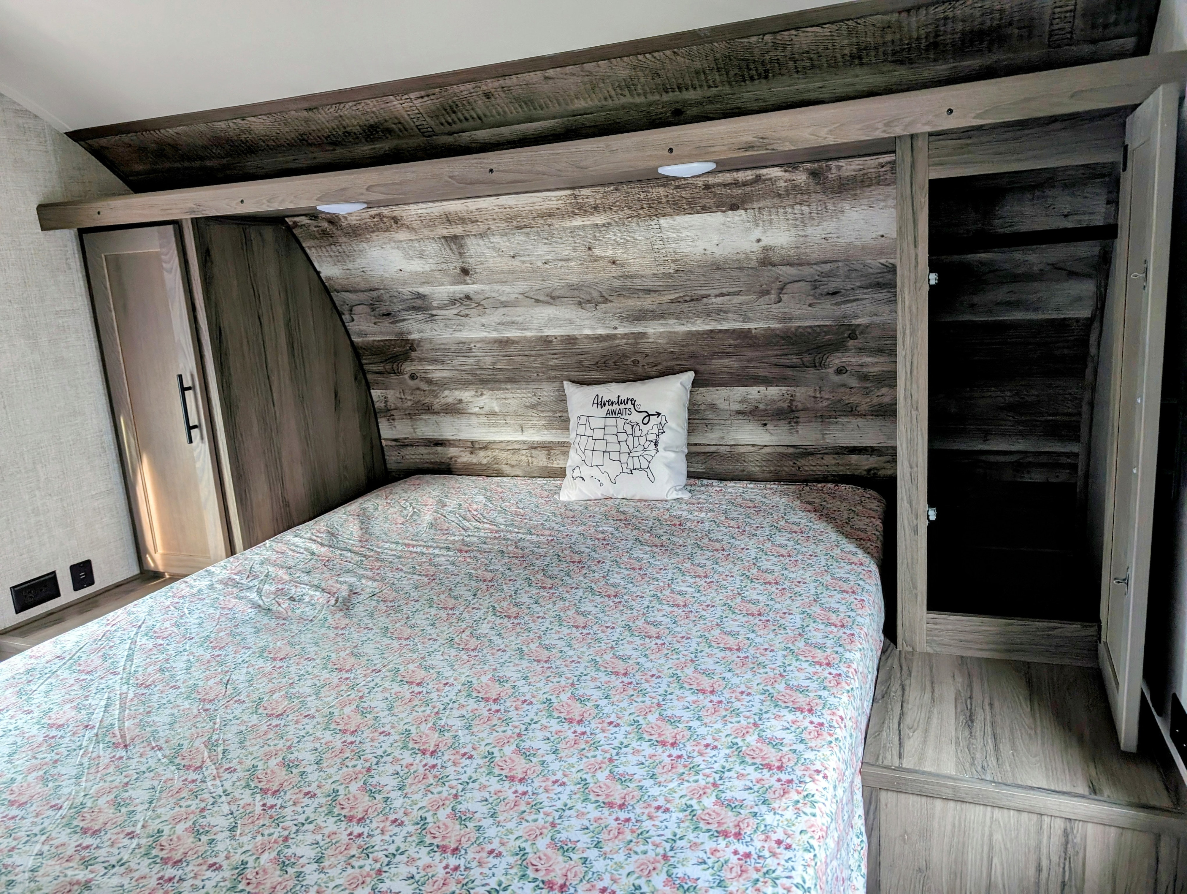 Cozy RV rental bedroom with rustic wood-look decor & storage. Features a bed with floral sheets & a travel-themed pillow.. Gulf Stream Ameri-Lite 2022