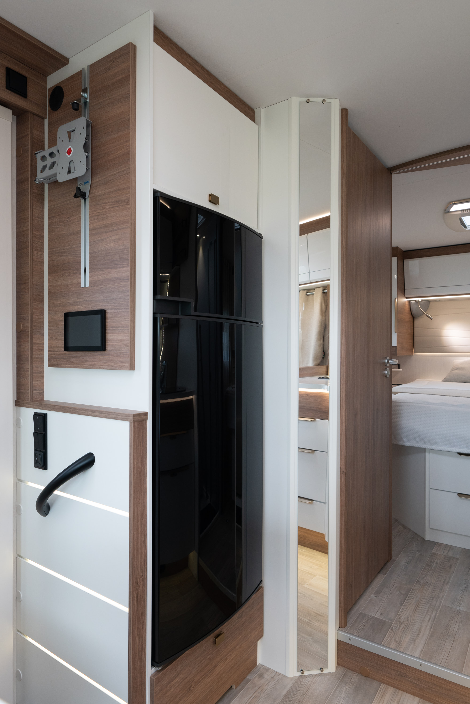 Modern RV rental interior: black refrigerator, TV mount, control panel, and entrance to a cozy bedroom area.. Pilote G740 FC EVIDENCE 2025
