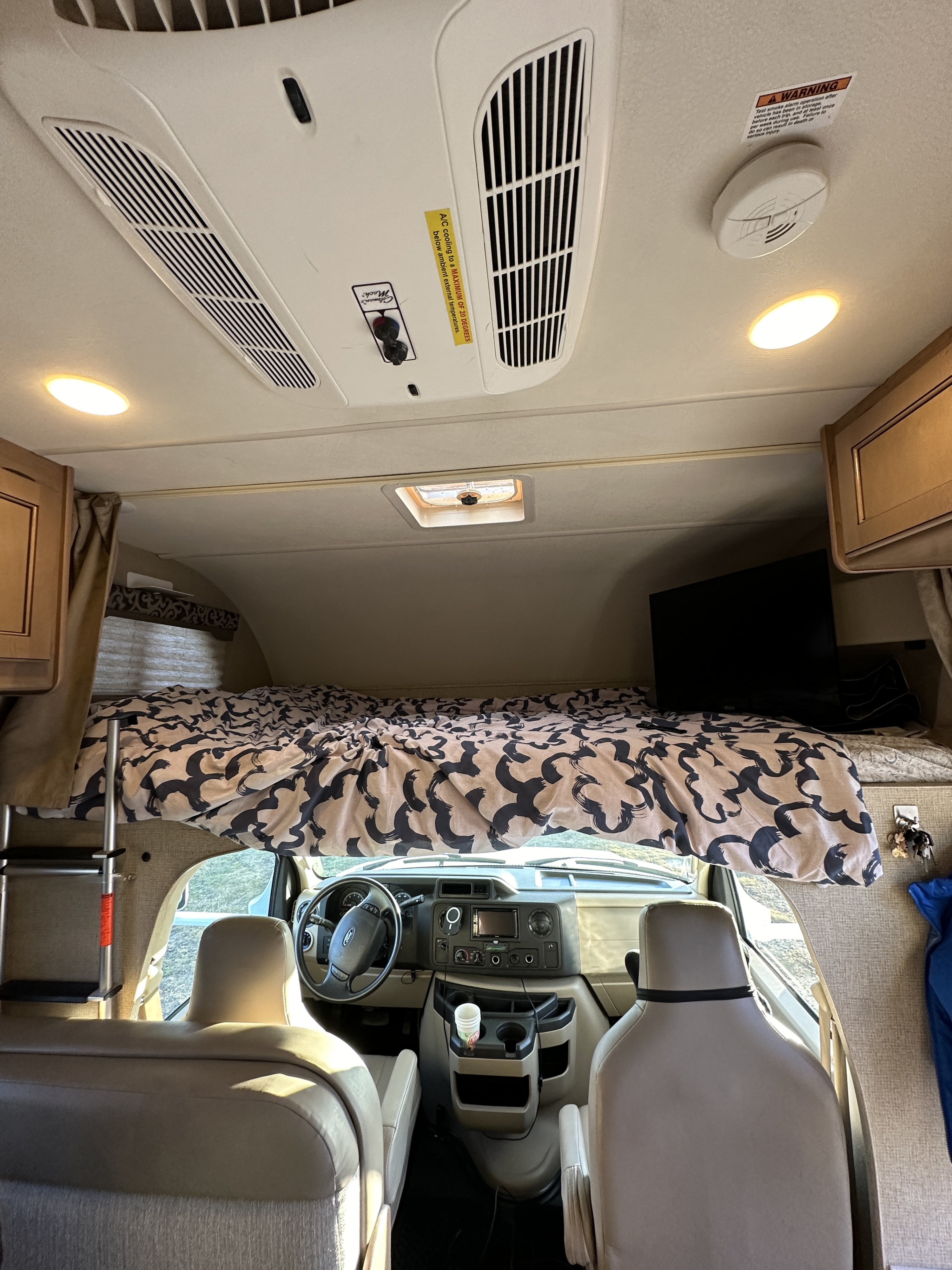 Inside an RV rental: cozy overhead bed, driver's area, and ceiling with AC vents, lights, and a smoke alarm visible.. Thor Four Winds 2019