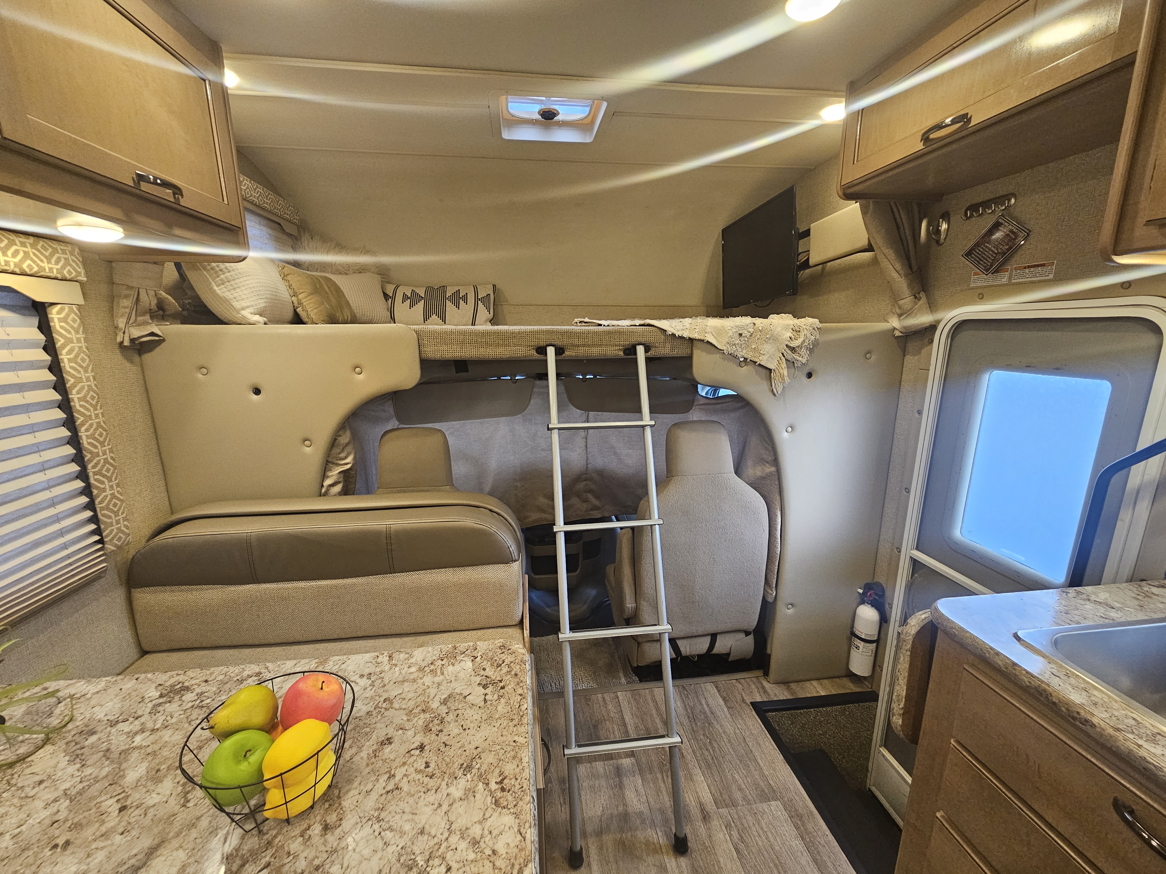 Cozy RV rental interior with dinette, kitchen, and over-cab bunk bed. Note the fire extinguisher by the door.. Thor Motor Coach Freedom Elite 2018