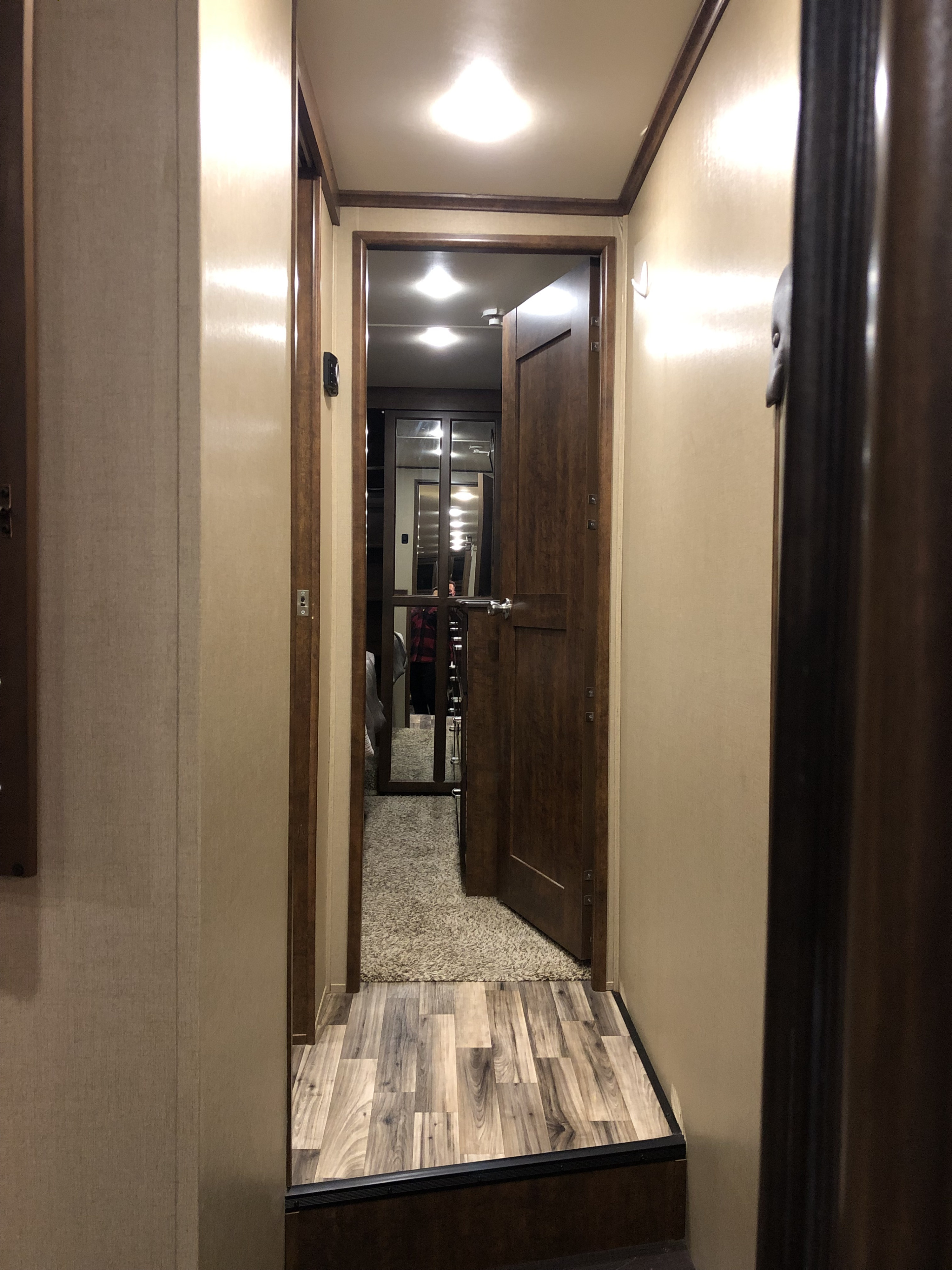 Inside an RV rental: a hallway with wood laminate transitioning to carpet. Dark wood trim frames multiple doors and bright ceiling lights.. Grand Design Solitude 2015