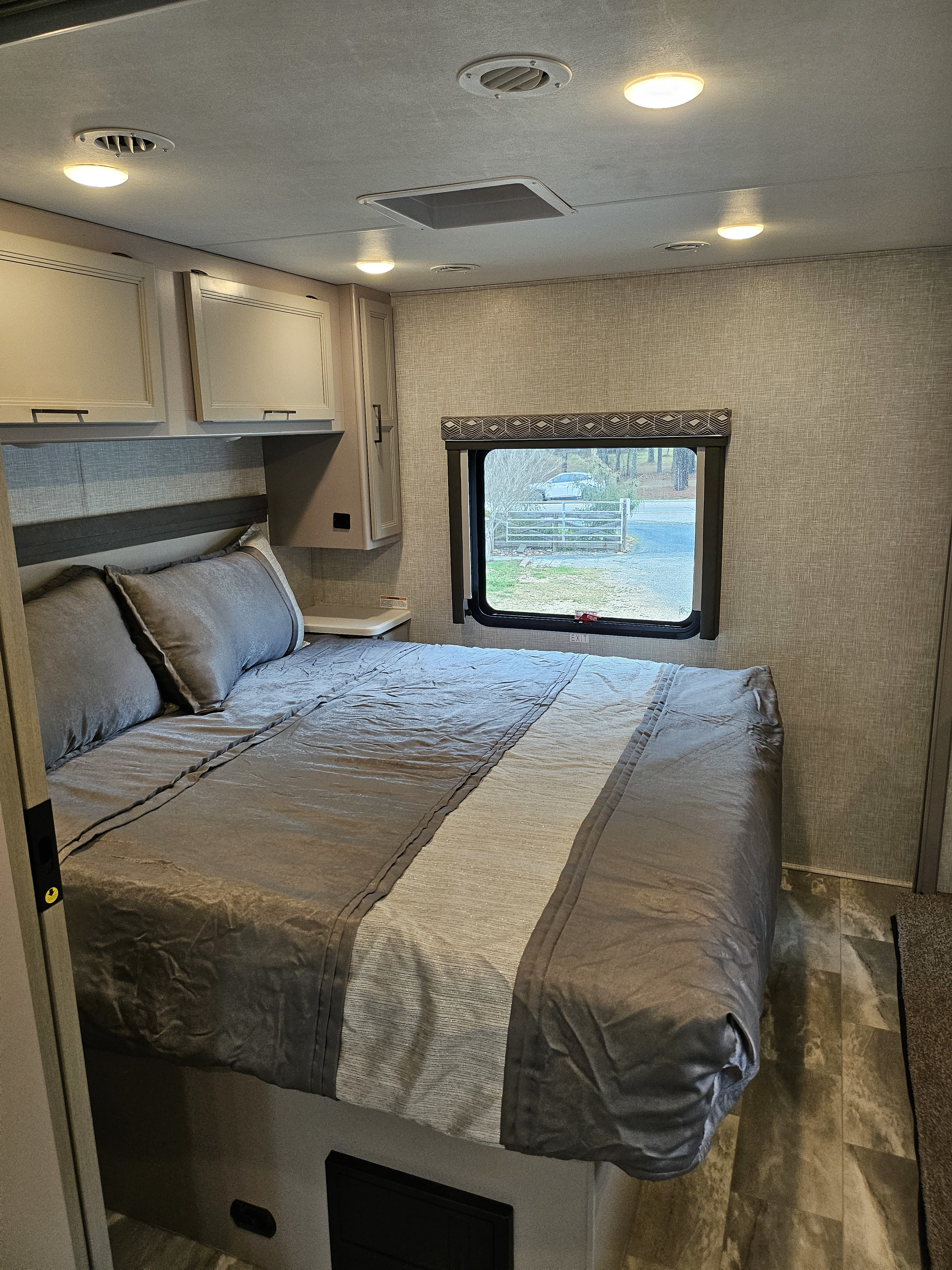 Spacious RV rental bedroom features a bed with gray bedding, overhead cabinets, and a window with outdoor views.. Thor Motor Coach Windsport 2022
