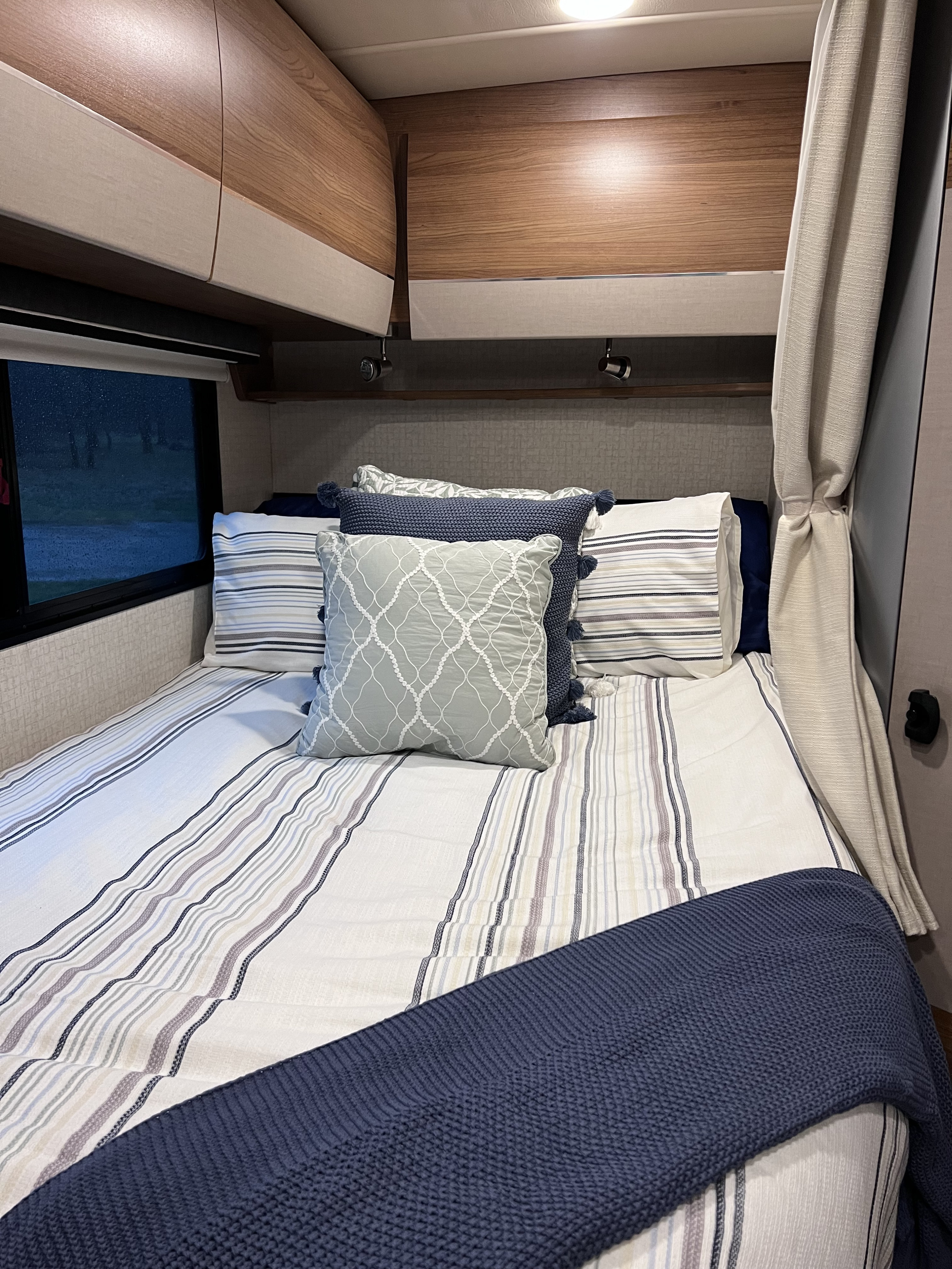 Cozy RV rental bedroom featuring striped bedding, throw pillows, and wood-toned overhead cabinets with a window view.. Winnebago Mercedes Navion 2016