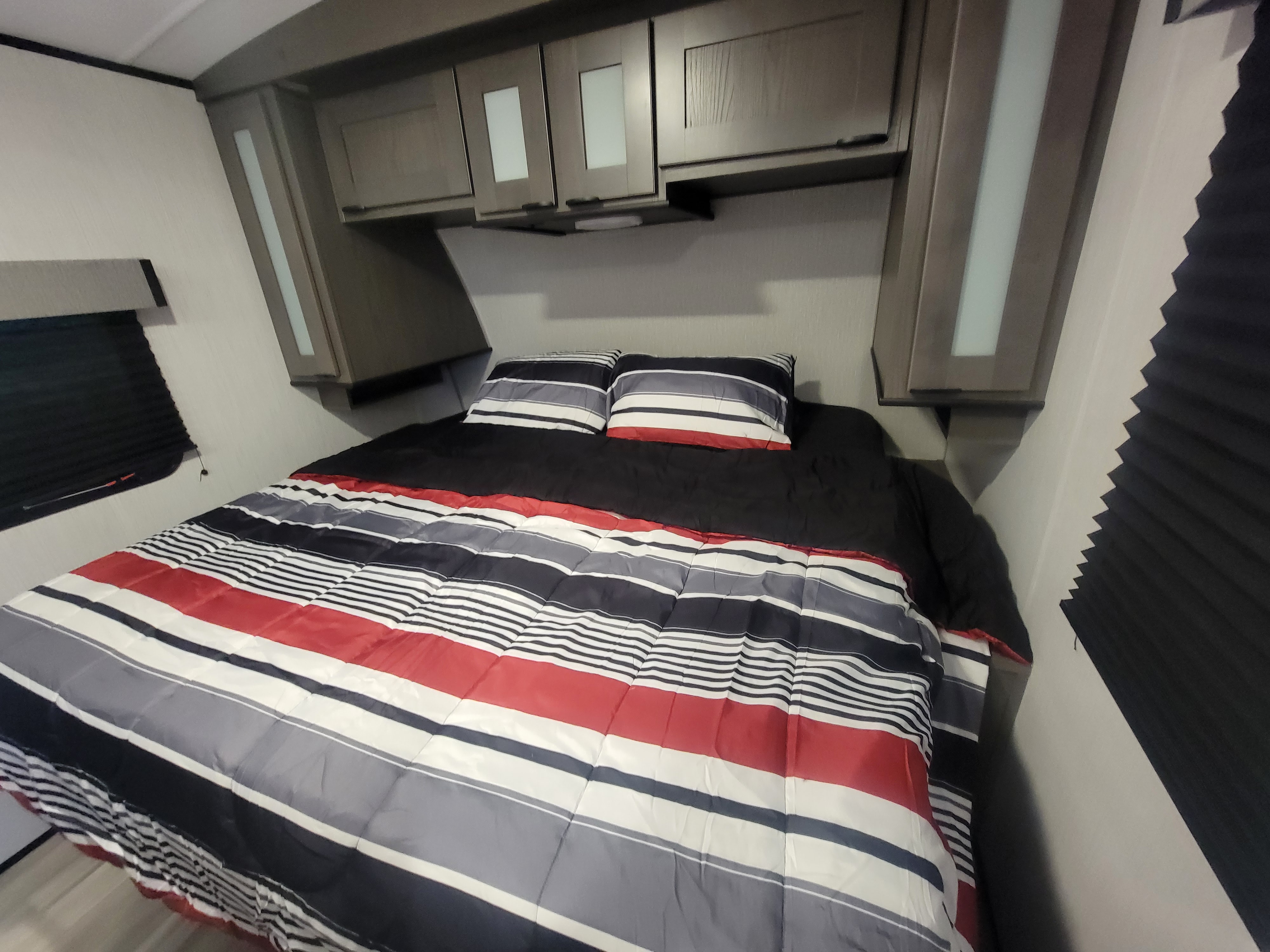 Cozy RV rental bedroom with a bed featuring red, white, black, and gray striped bedding, overhead cabinets, and window blinds.. Cruiser Rv Corp MPG 31BH 2023