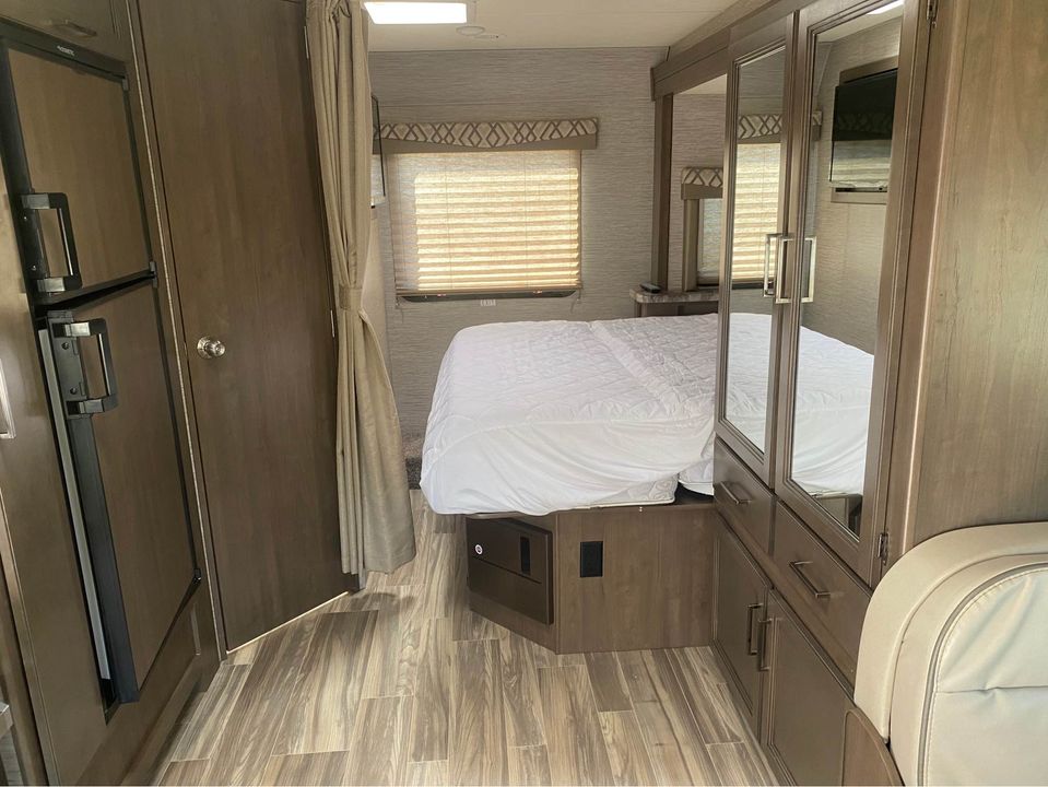Cozy RV rental bedroom with white bed, mirrored wardrobe, and TV. Features refrigerator, door, and wood-look flooring.. Thor Four Winds 2021