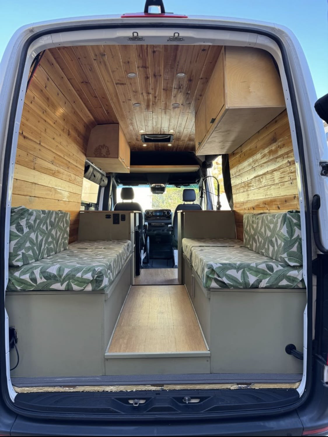 Spacious RV rental interior from the rear: wooden ceiling/walls, leaf-patterned benches, central aisle, and storage.. Mercedes-Benz Sprinter 419 LWB High Roof 2019