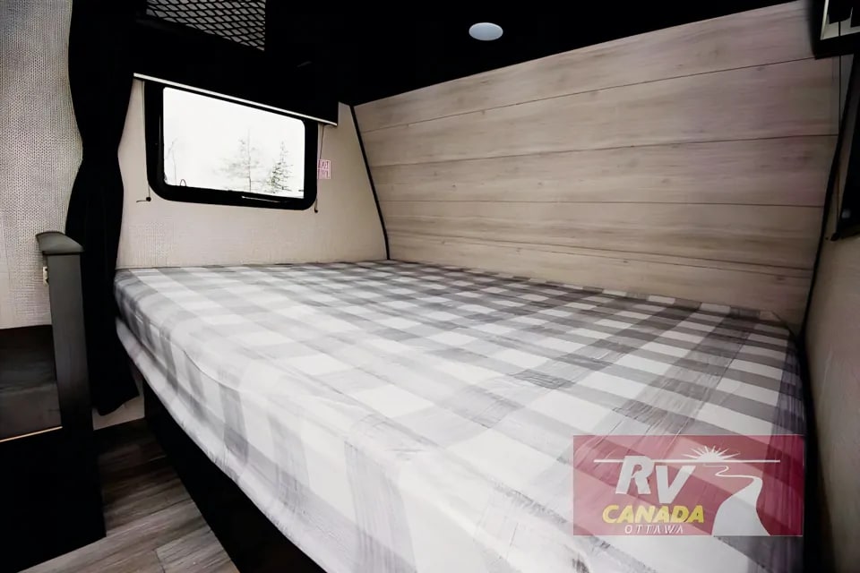 RV rental interior: a comfy bed with grey plaid sheets, a window showing trees, and light wood paneling.. Jayco Jay Flight SLX 2023
