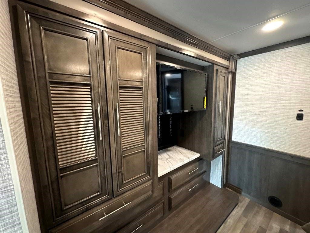 RV rental interior: dark wood cabinetry with louvered doors, a mounted TV, and dresser drawers for storage.. Entegra Coach Vision XL 2024
