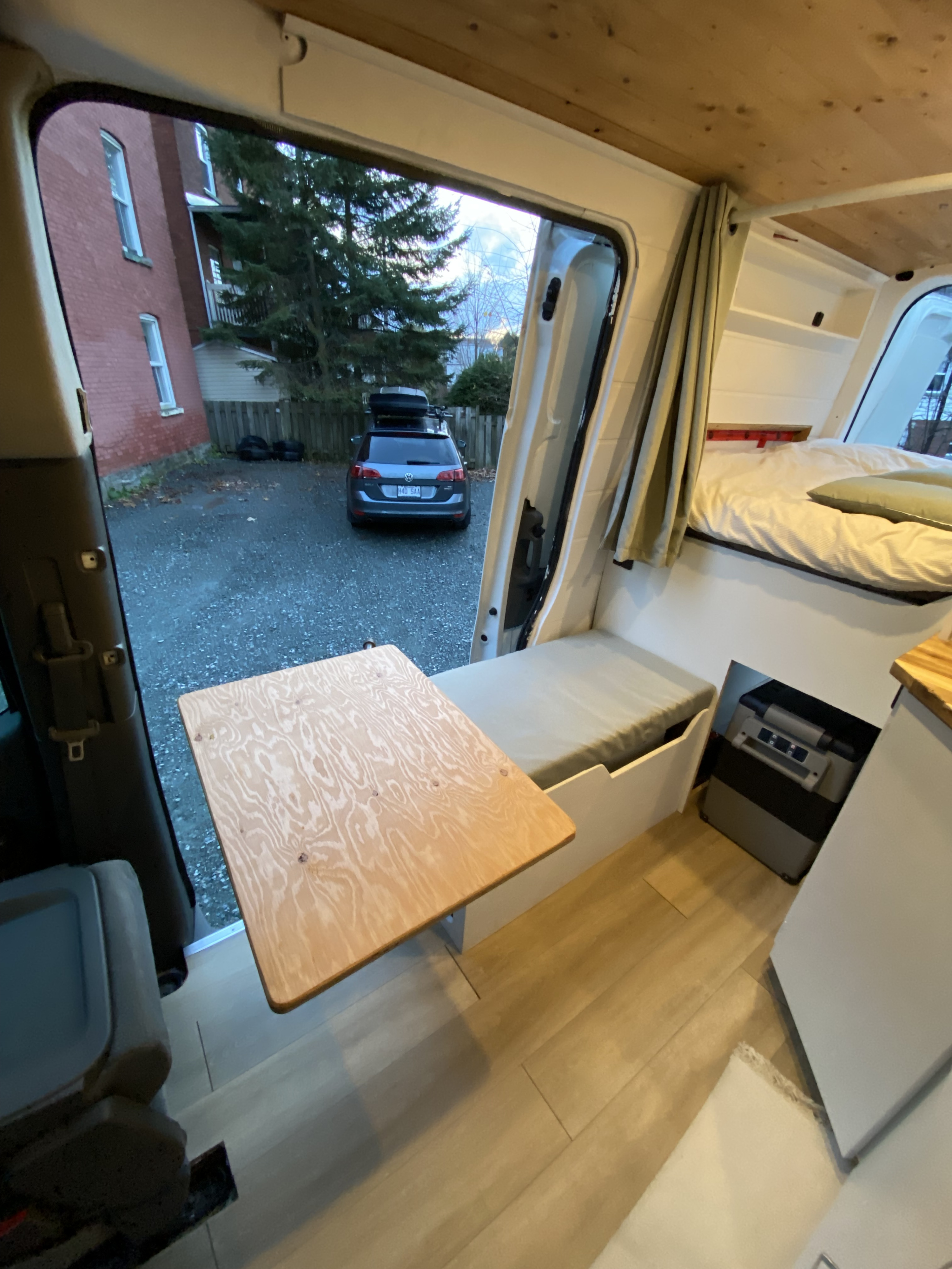 Cozy RV rental camper van interior. Features a custom wooden table, comfy bed, and cooler. Ready for adventures!. Nissan PRIMASTAR 2012