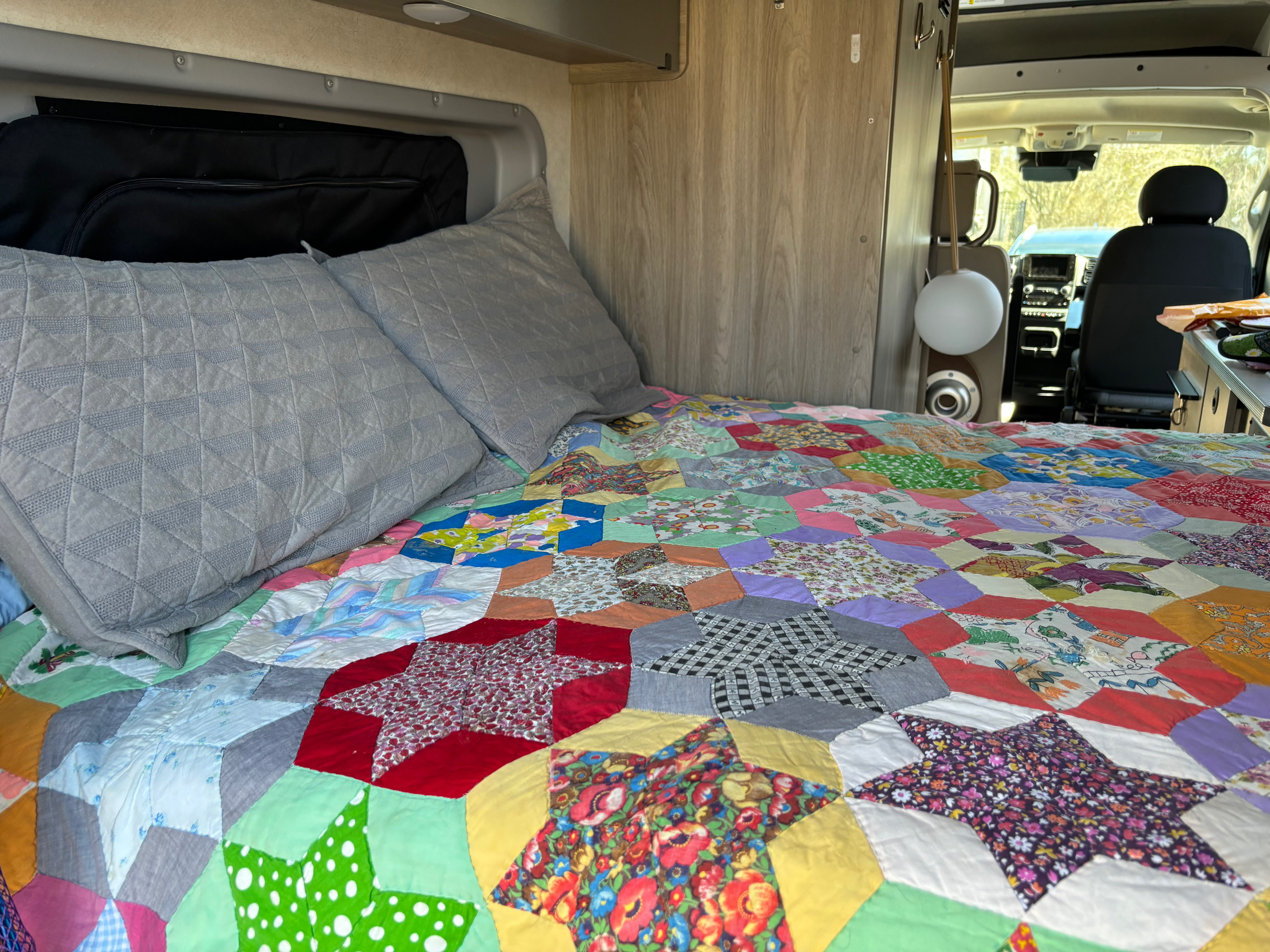 Cozy rv rental bed with a vibrant patchwork quilt & grey pillows. Features a view into the cockpit.. Winnebago Solis 2023