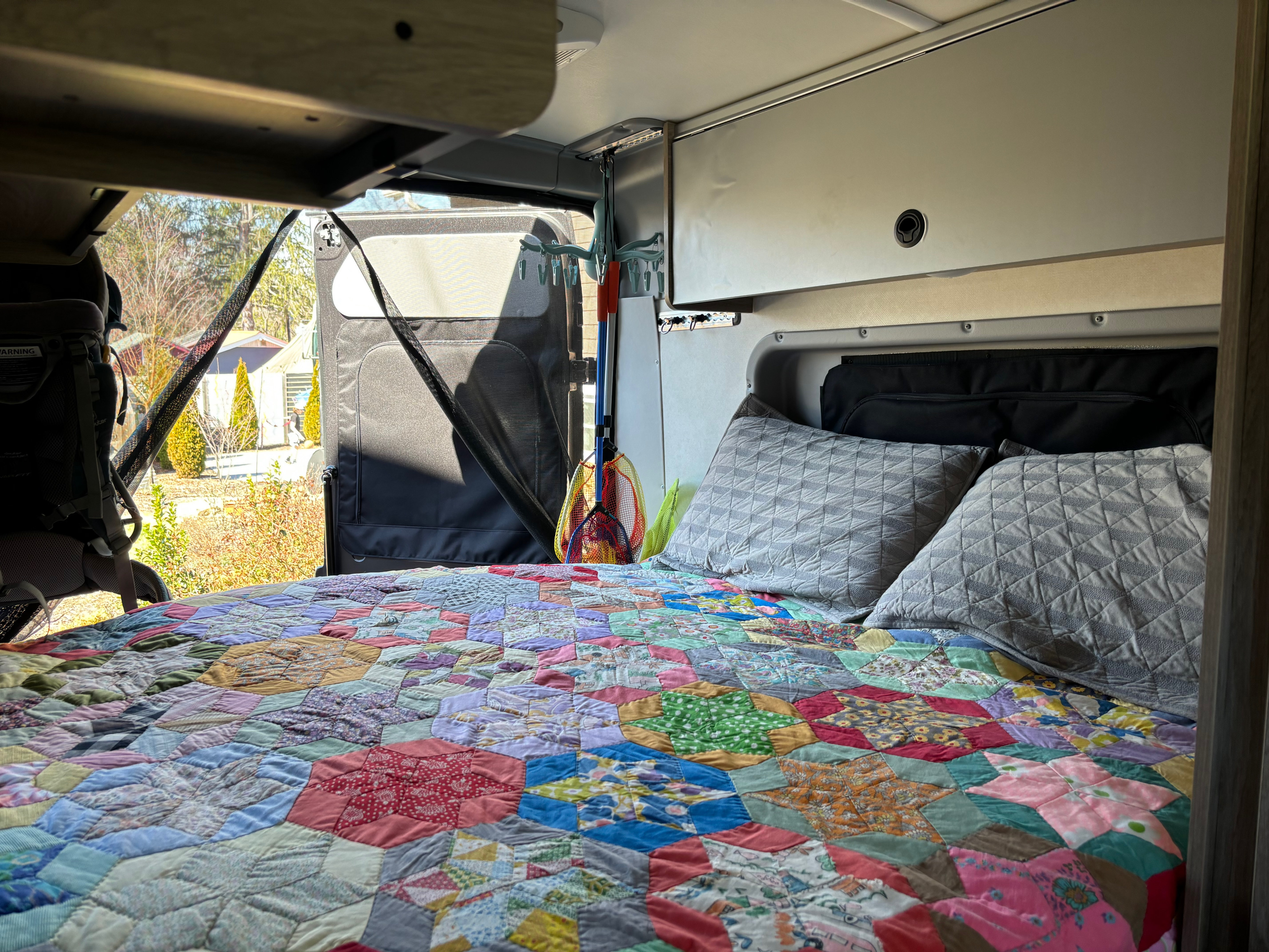 Cozy rv rental bedroom with a colorful patchwork quilt on the bed, looking out to trees and houses through an open screened door.. Winnebago Solis 2023