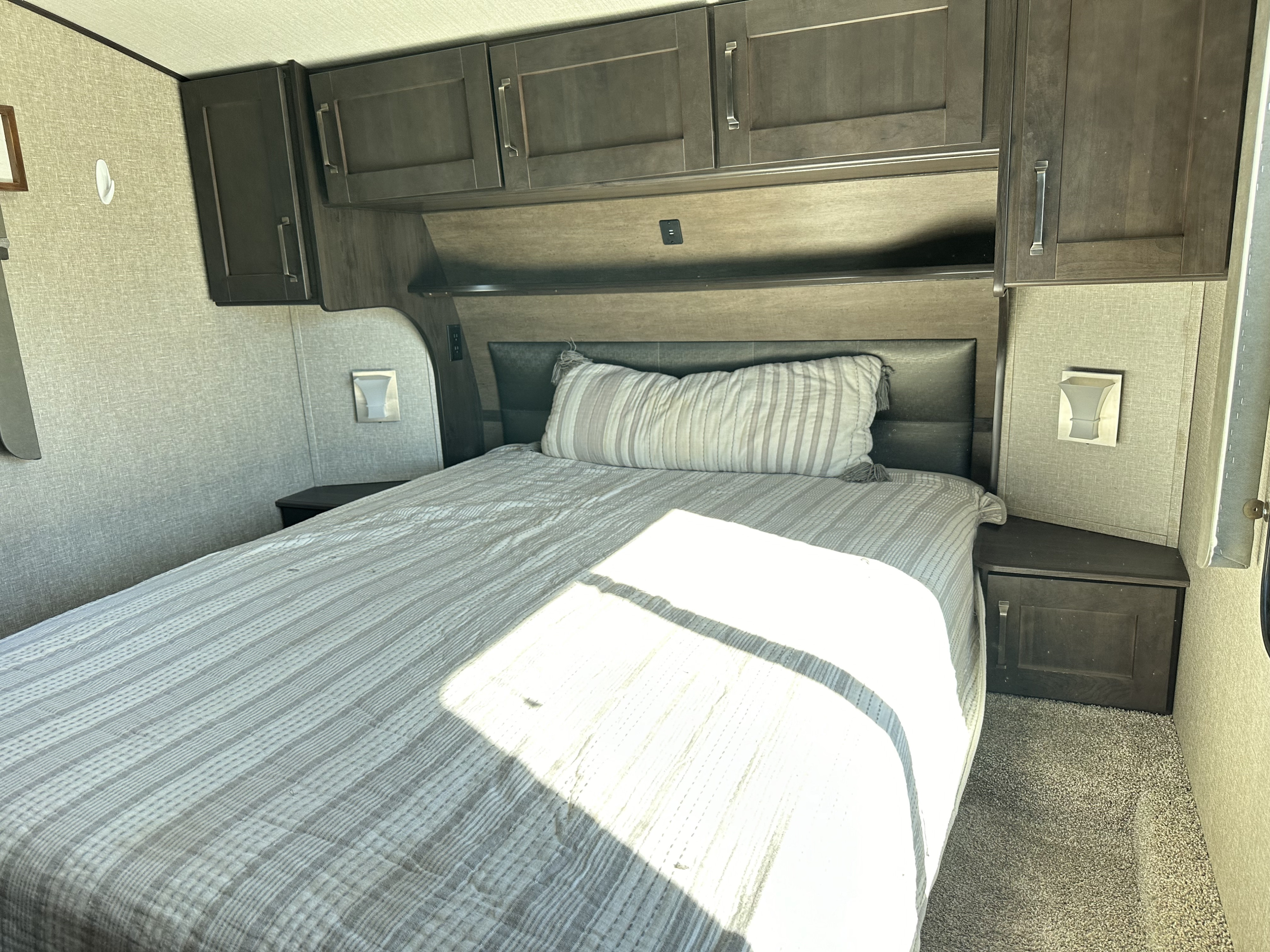Comfortable RV rental bedroom featuring overhead cabinets, a striped bedspread, and nightstands with integrated lighting.. Grand Design Reflection 2021