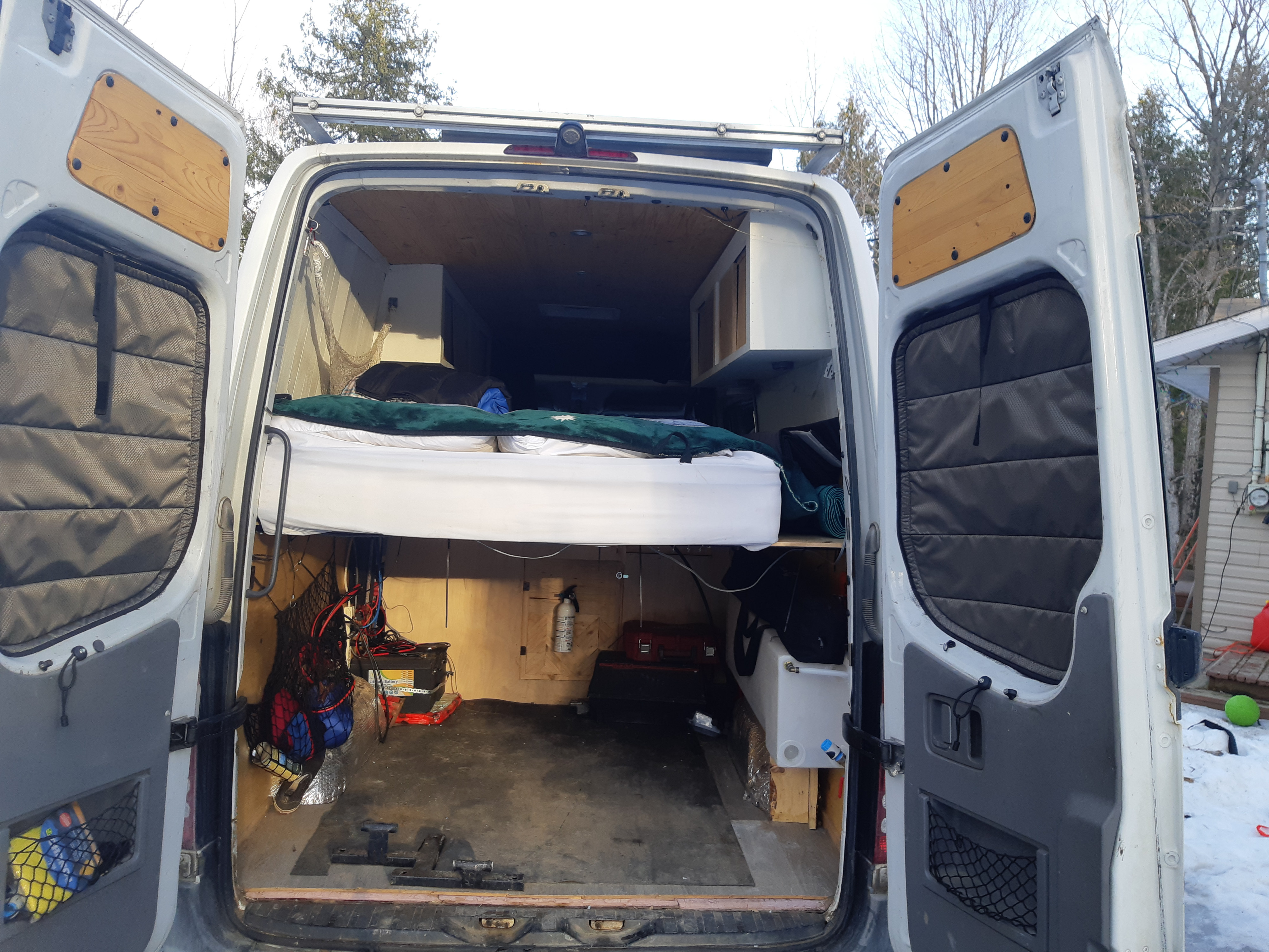 Open rear doors of an RV rental camper van conversion, showing elevated bed, under-bed storage, and utility setup.. Dodge Sprinter 2007