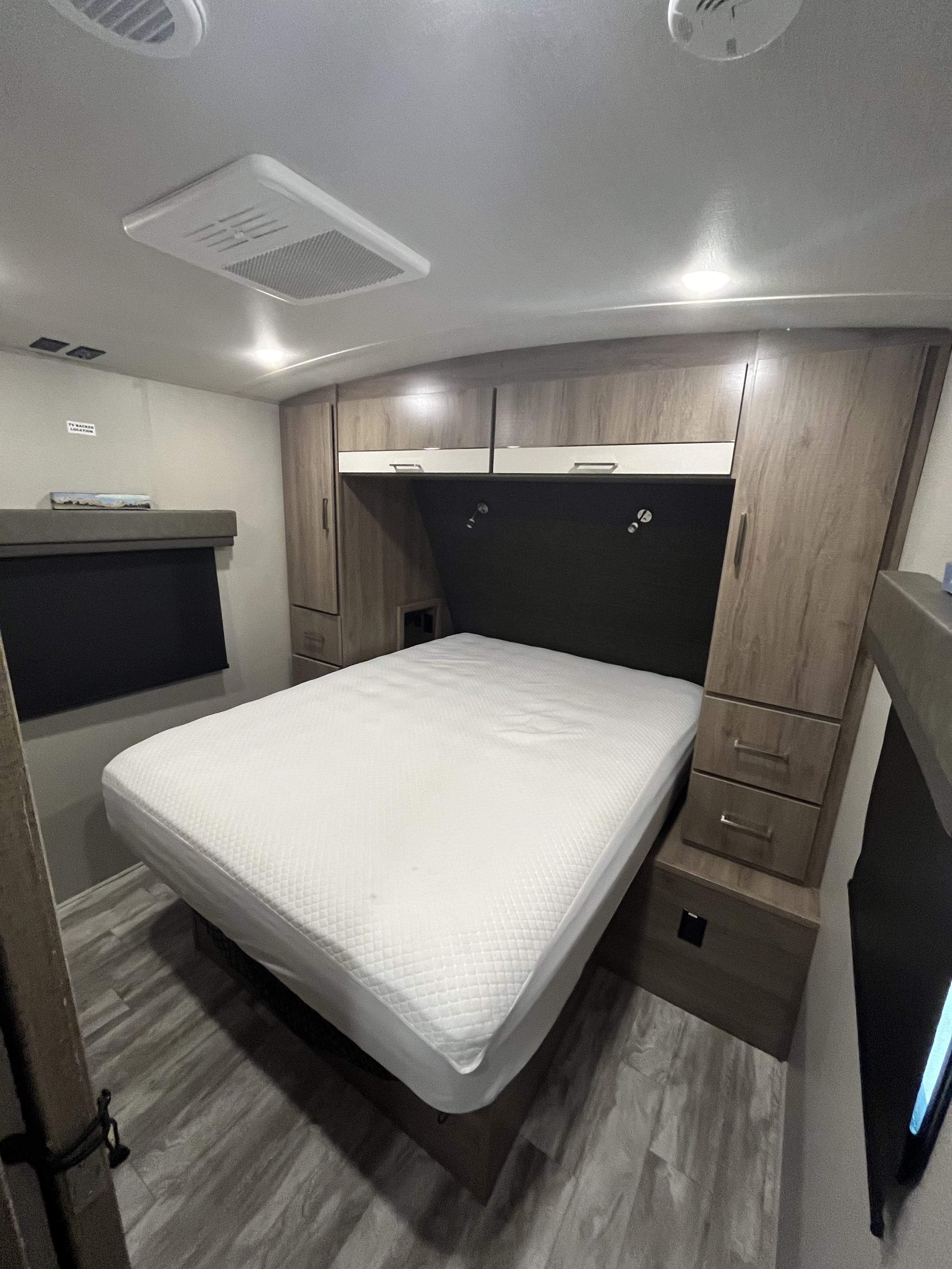 This RV rental bedroom features a white bed, wood-grain cabinets, overhead AC, and comfortable lighting.. Grand Design Imagine 2023