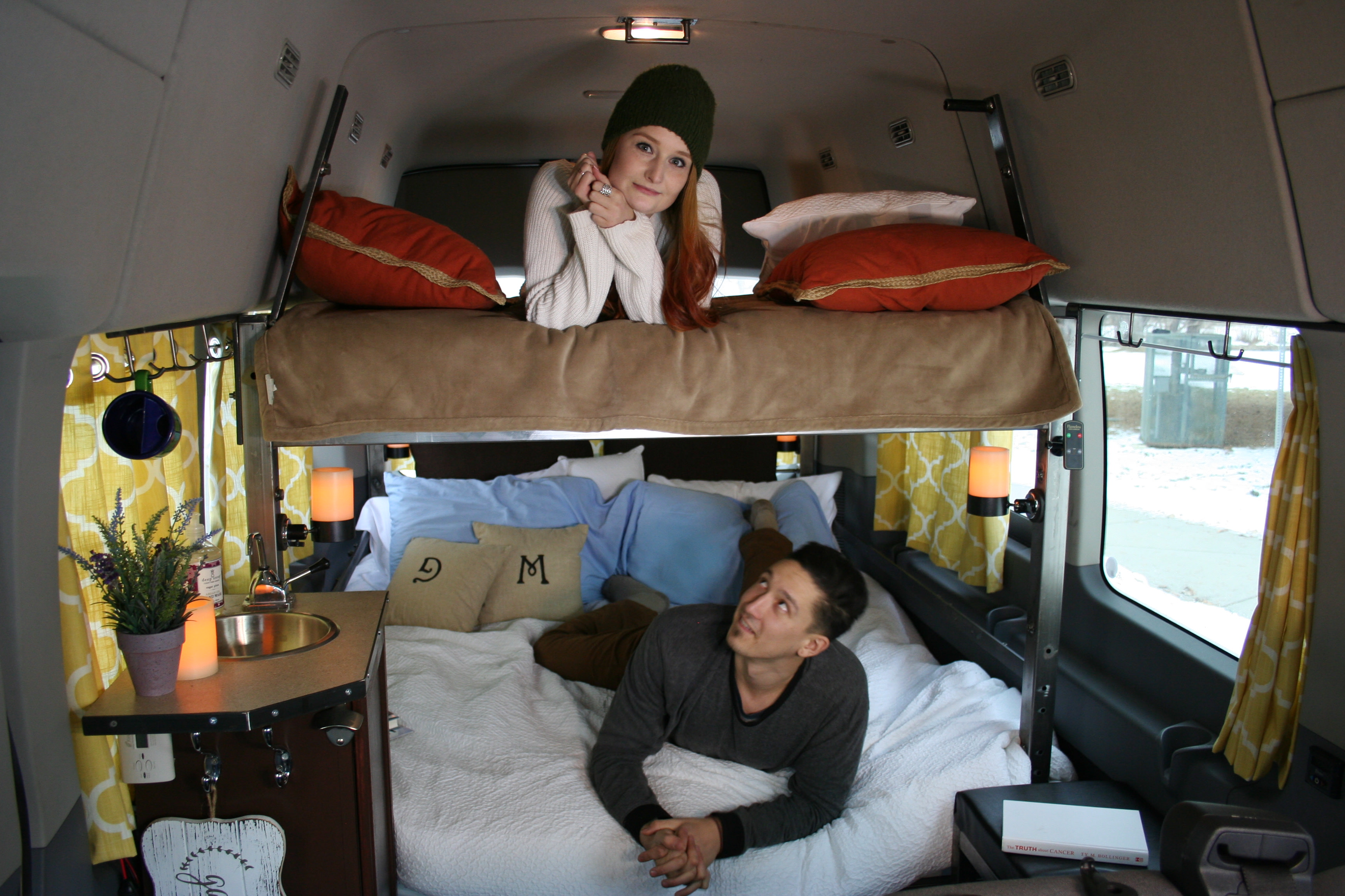 Cozy RV rental interior shows a woman on an upper bunk and a man on the lower bed inside a custom camper van.. Ford Transit Custom 2023