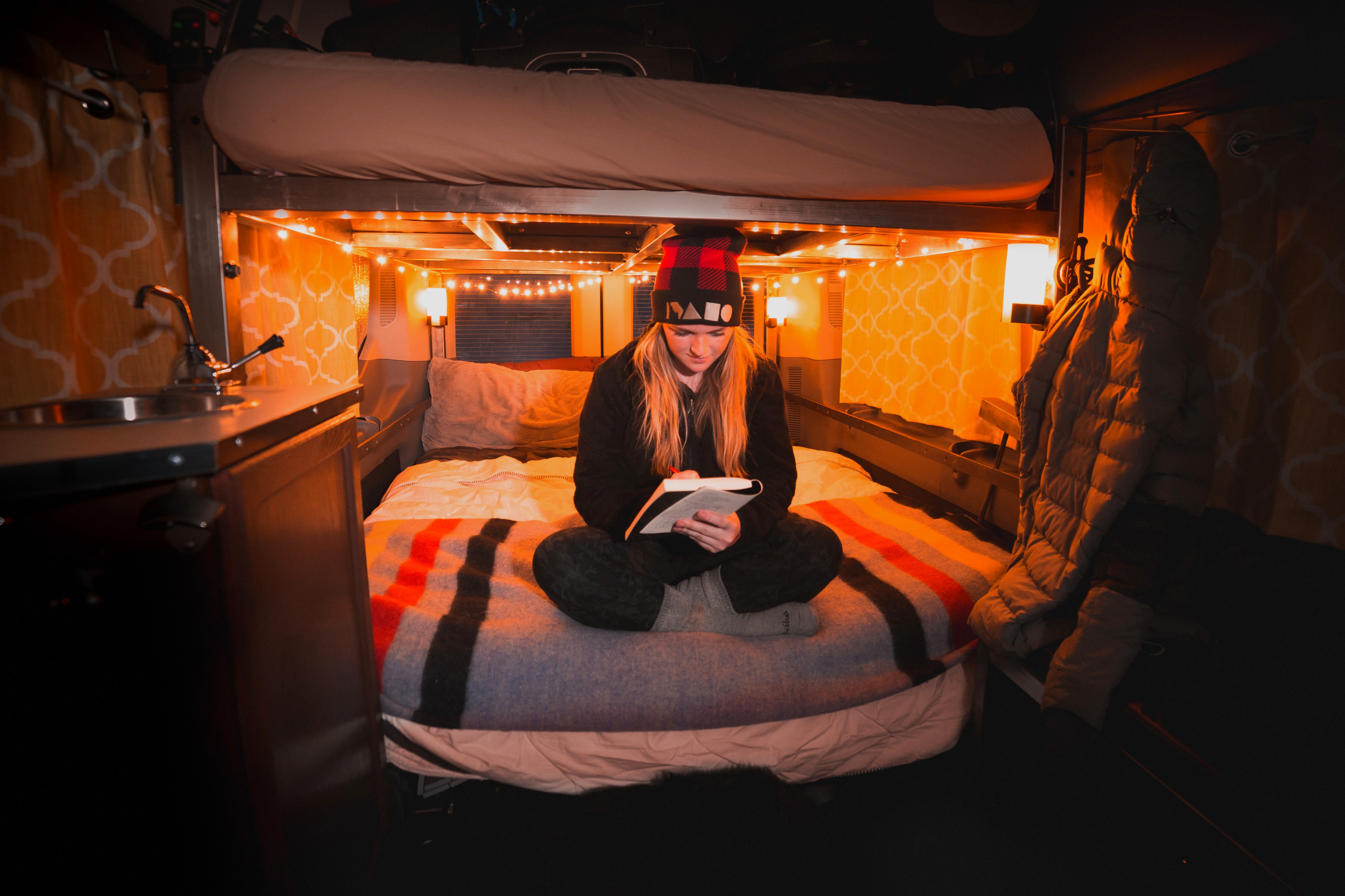 Cozy RV rental interior: A woman reads on a comfortable bed under warm string lights, featuring a bunk bed above and small sink area.. Ford Transit Custom 2023