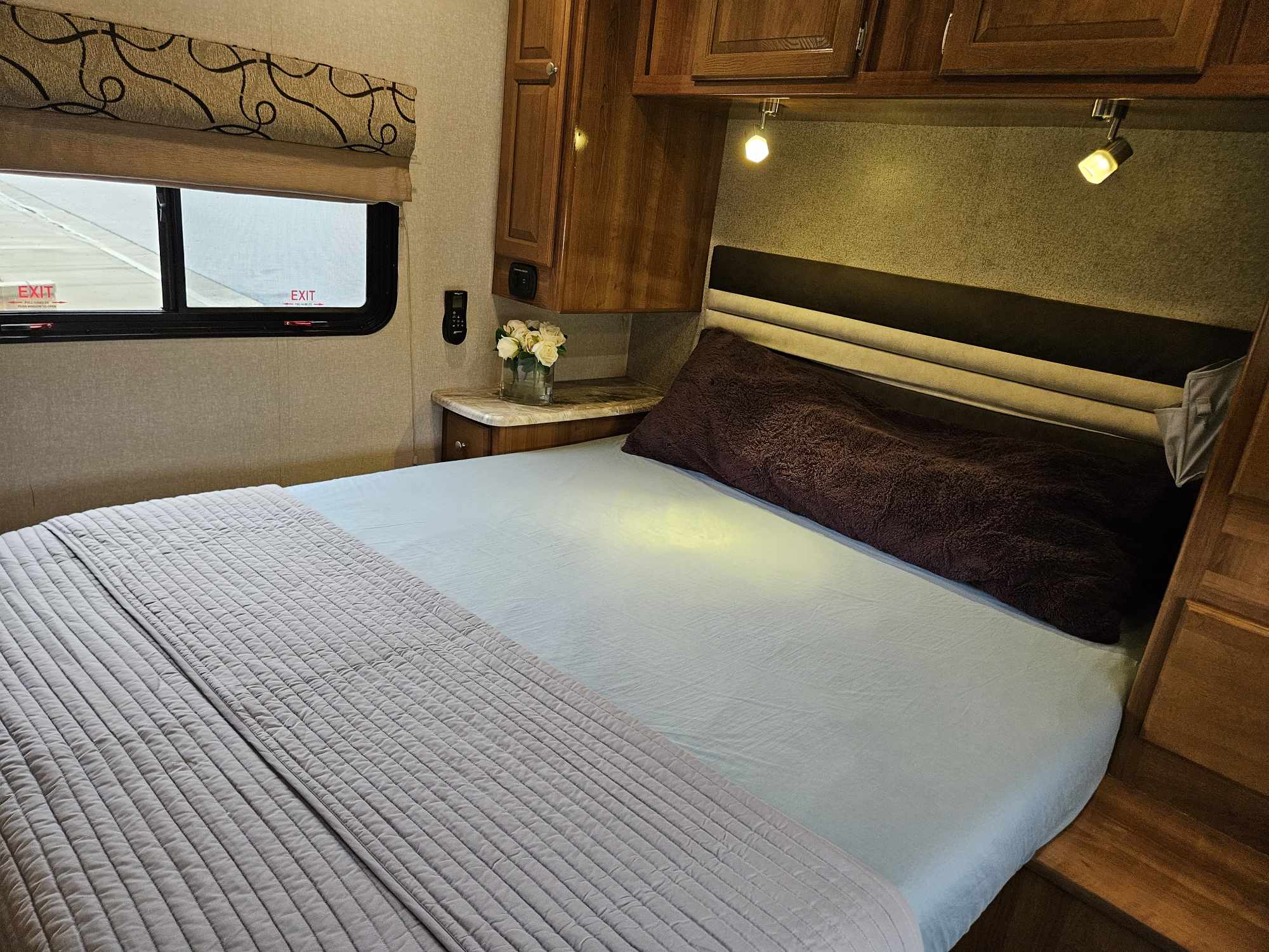 Comfortable RV rental bedroom: light blue bed, brown pillow, wood cabinets, and emergency exit windows.. Coachmen Leprechaun 2020