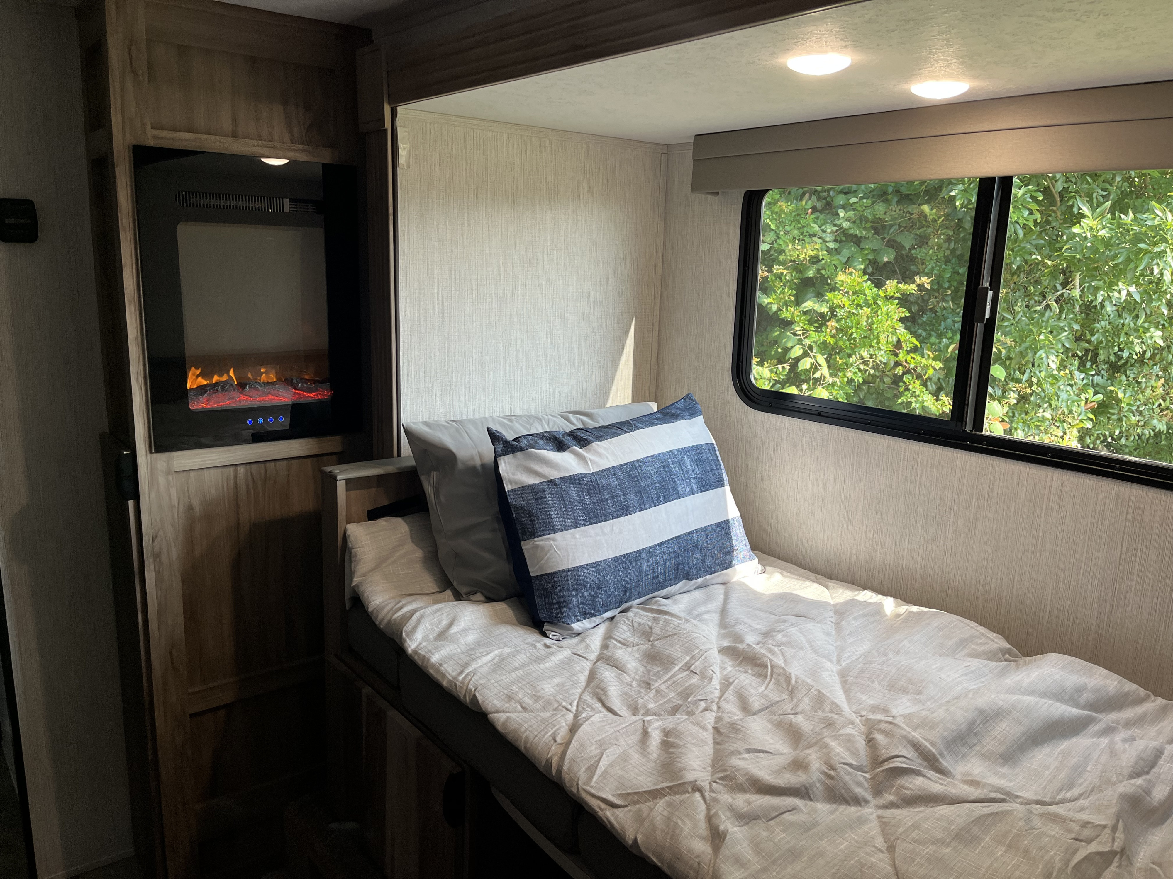Inside a cozy RV rental: a comfortable bunk with a striped pillow, a fireplace, and a window framing lush trees.. Palomino Palomini 2022