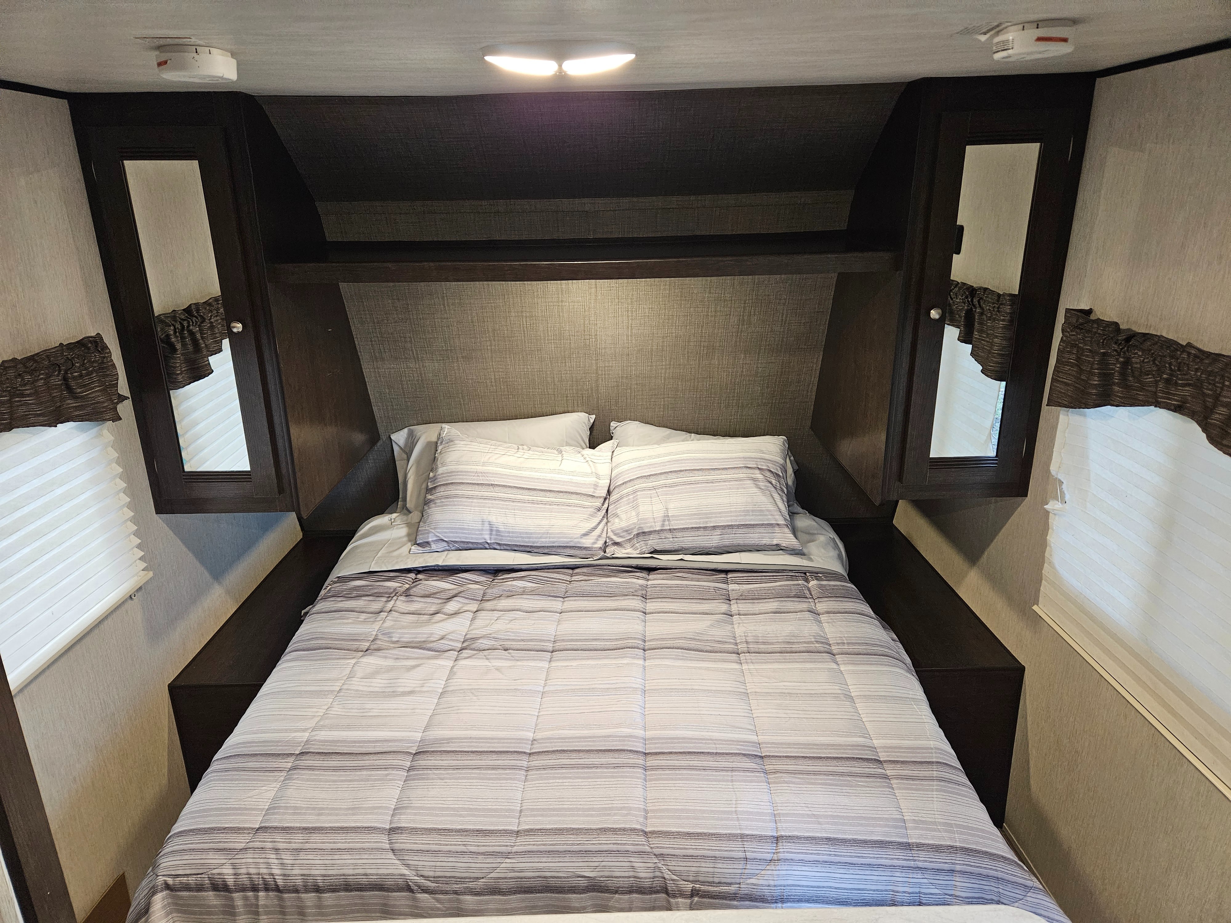 Cozy RV rental bedroom featuring a full bed with striped comforter, pillows, side windows, and overhead storage.. Heartland RVs Trail Runner SLE 2019