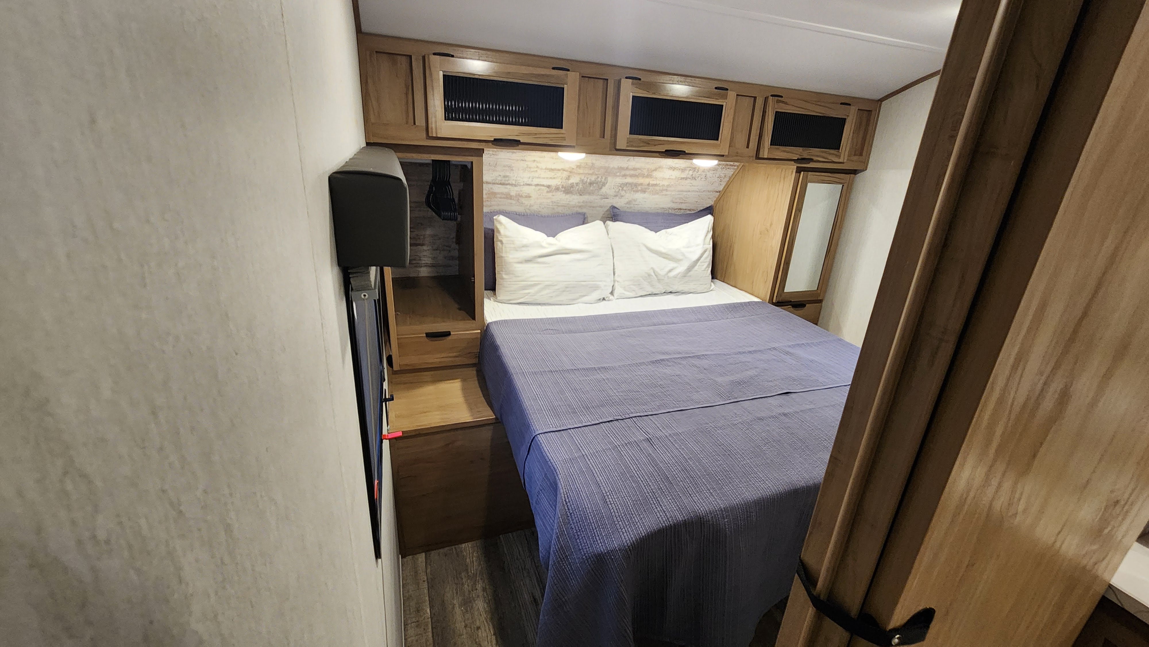 Inside this RV rental, find a cozy bedroom with a comfortable bed, overhead wood cabinets, and a wall-mounted TV.. Forest River 22sw-l 2024