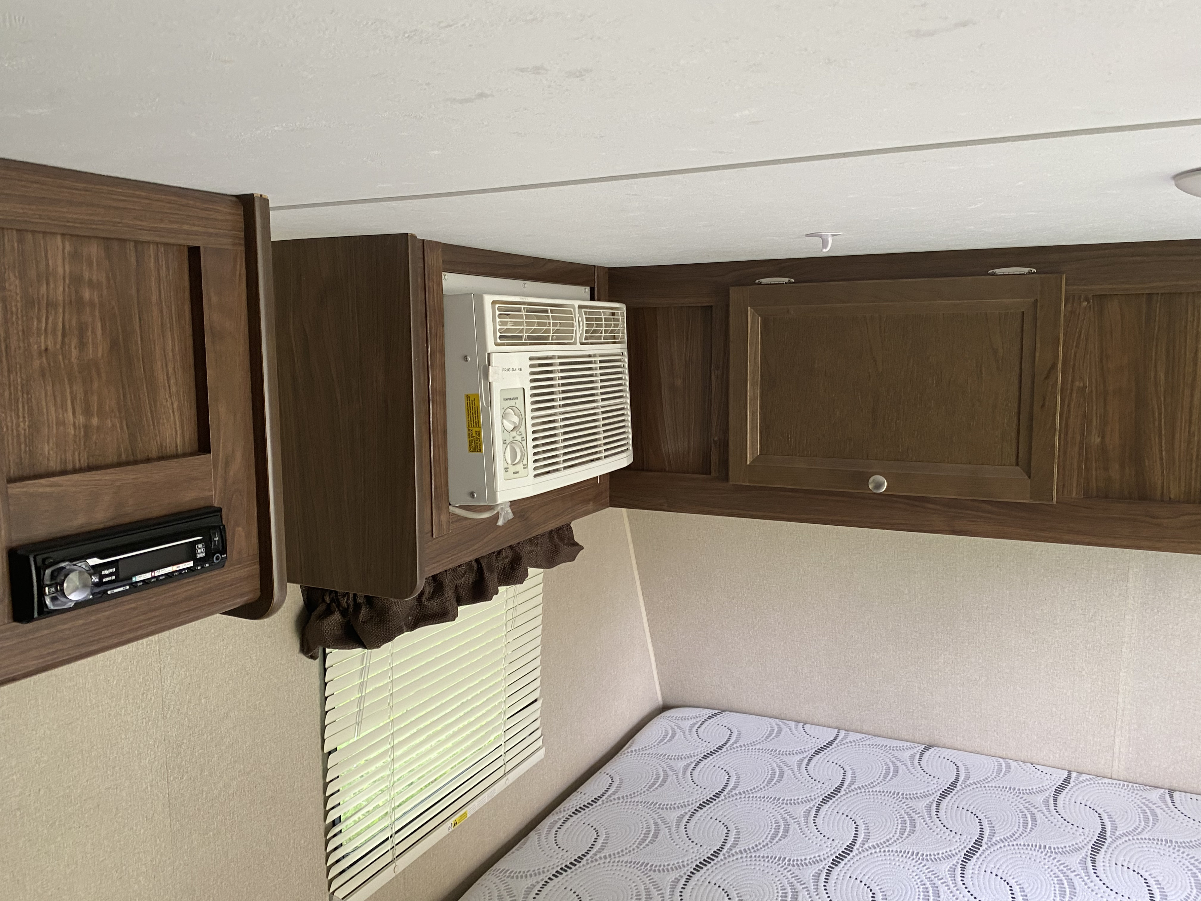 Inside an RV rental: a compact sleeping area with built-in wooden cabinets, a window AC, stereo, and a bed.. Dutchmen Coleman Lantern 2016