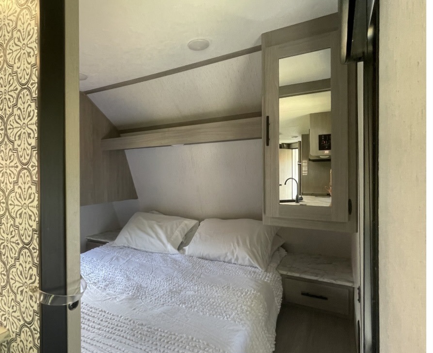 RV rental bedroom features a comfy bed, white linens, storage, and a mirrored cabinet reflecting the kitchen space.. Coleman Dutchman 2021