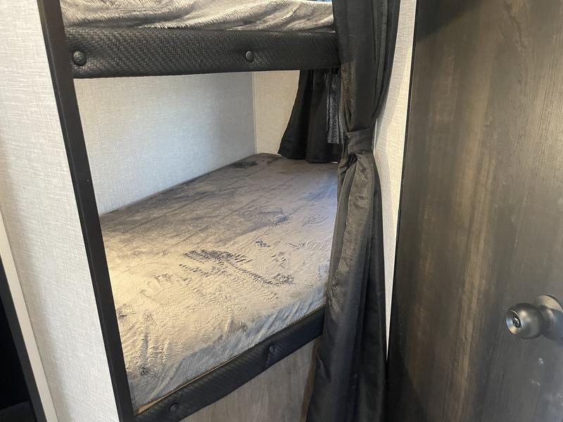 Interior of an RV rental: two compact bunk beds with grey bedding & dark frames. A tied-back privacy curtain is near a wooden door.. Jayco Jay Flight 235 MBHW 2024