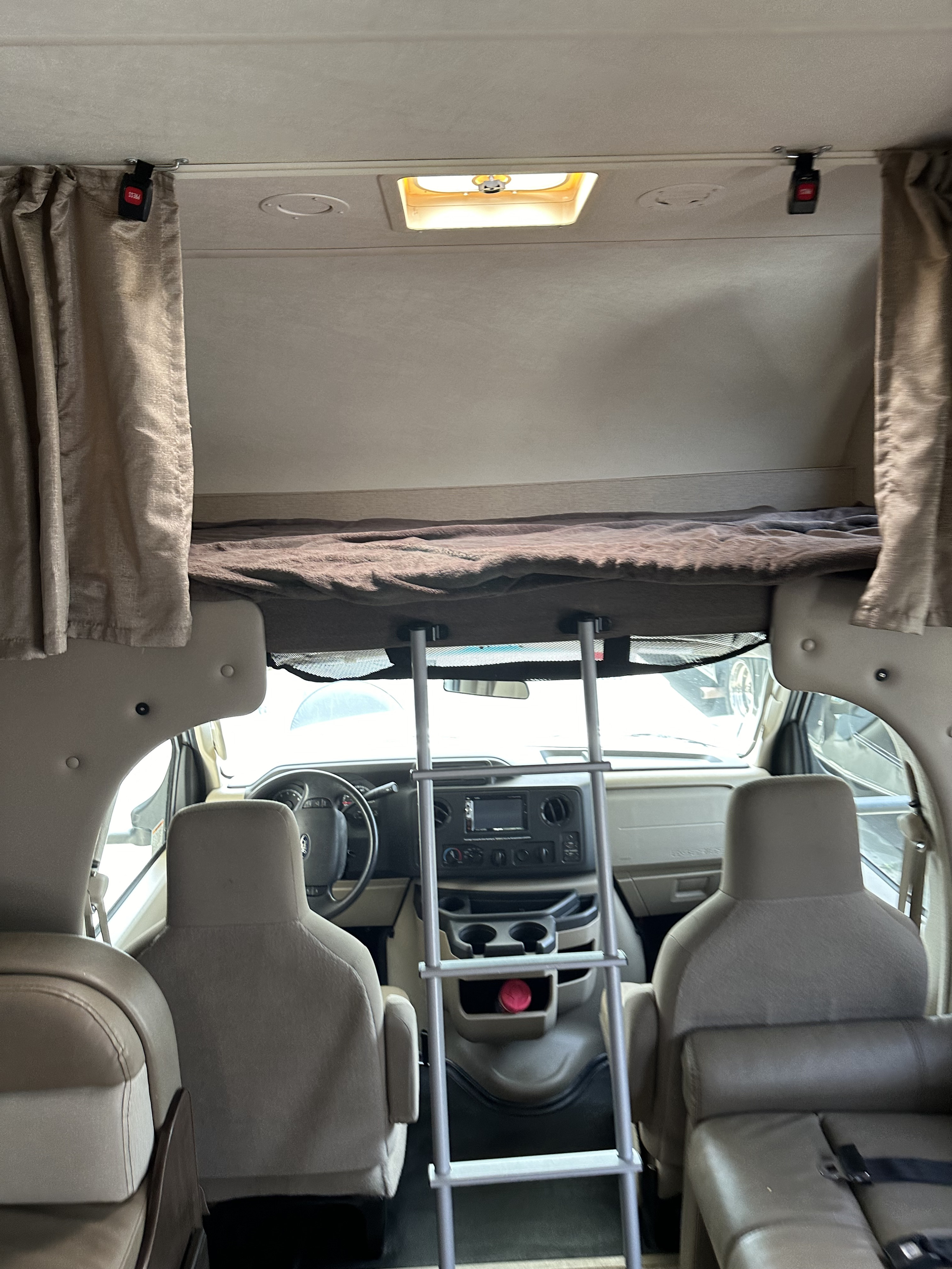 An rv rental's interior features an overhead bunk bed with a ladder leading to the driver's cockpit and dashboard.. Thor Motor Coach Four Winds 2020