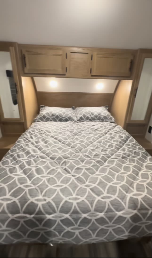 This RV rental bedroom has a gray geometric patterned bed, overhead cabinets, and bedside storage.. Cherokee Grey Wolf 2023