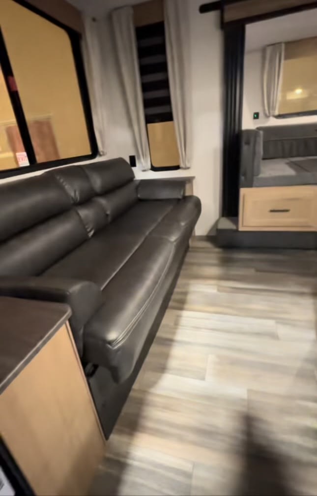 This RV rental features a comfortable living space with a long black leather sofa, large windows, and a glimpse of the sleeping area beyond.. Cherokee Grey Wolf 2023