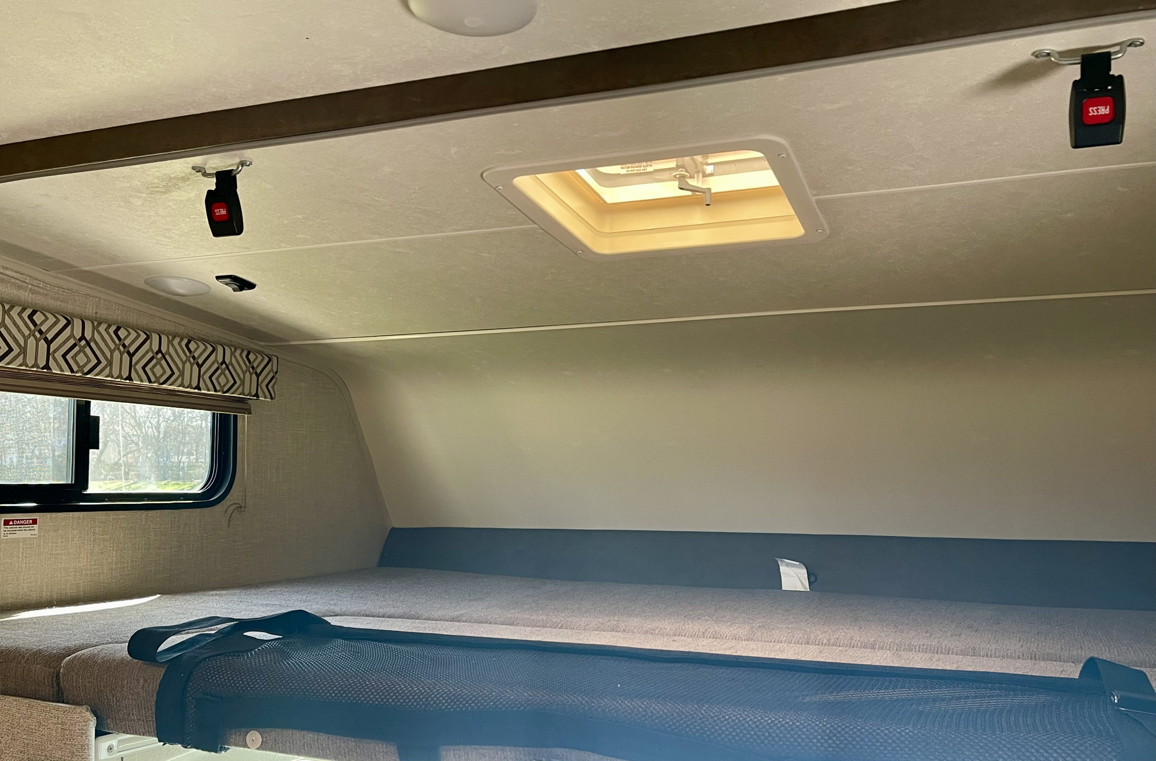 An RV rental's cabover bunk area, featuring a bed with a safety net, window, ceiling vent, and seatbelt buckles.. Forest River Coachmen Leprechaun 2020