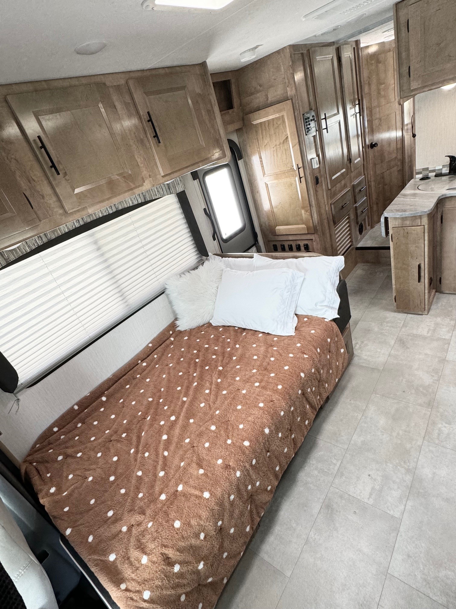 Cozy RV rental interior featuring a bed with a brown polka-dot blanket, wood cabinets, and a kitchen area.. Forest River Coachmen Leprechaun Premier 2021