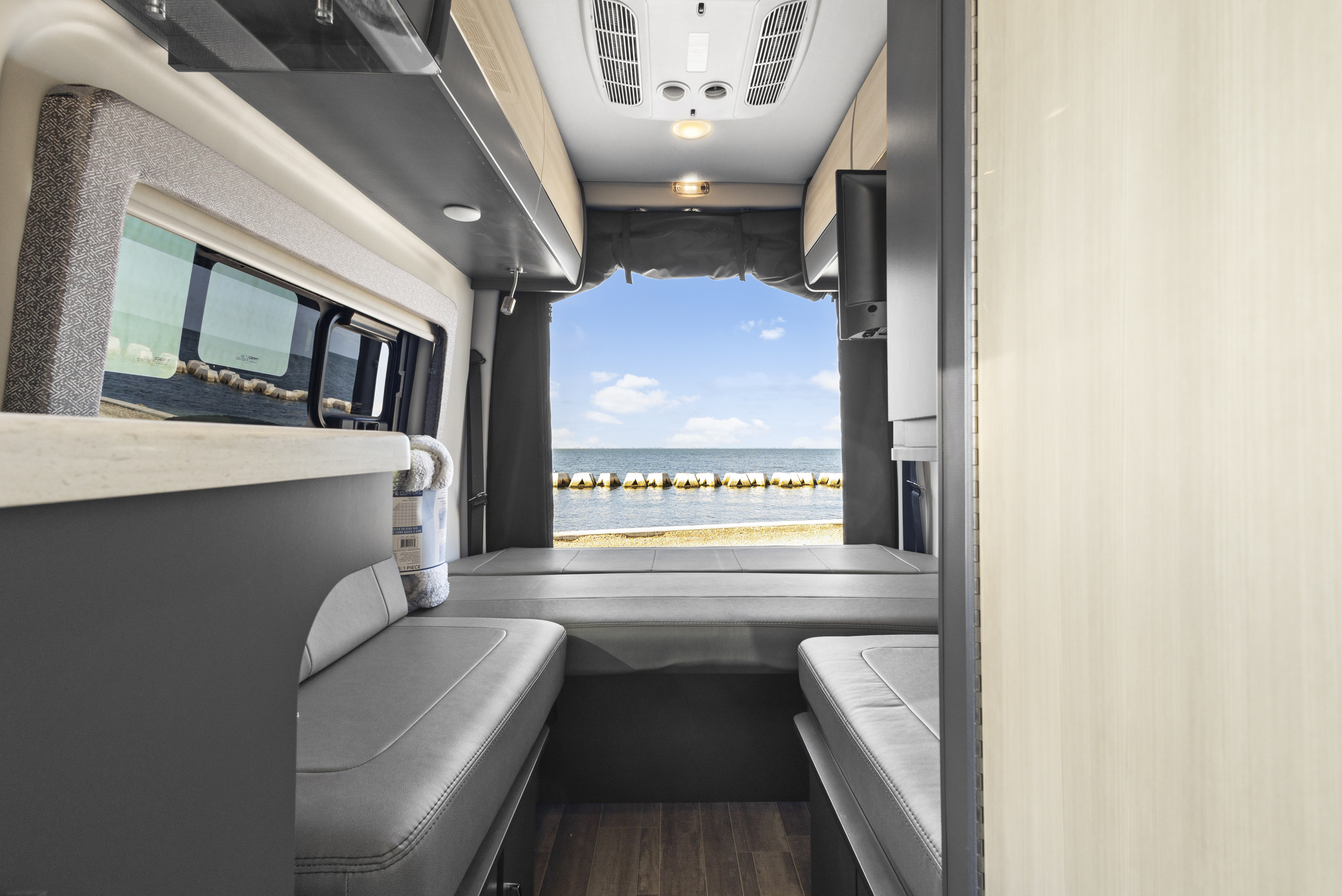 Step inside this RV rental! View of a modern camper van interior, featuring grey seating, wood-grain accents, and a large ocean view.. Thor Sanctuary 2023