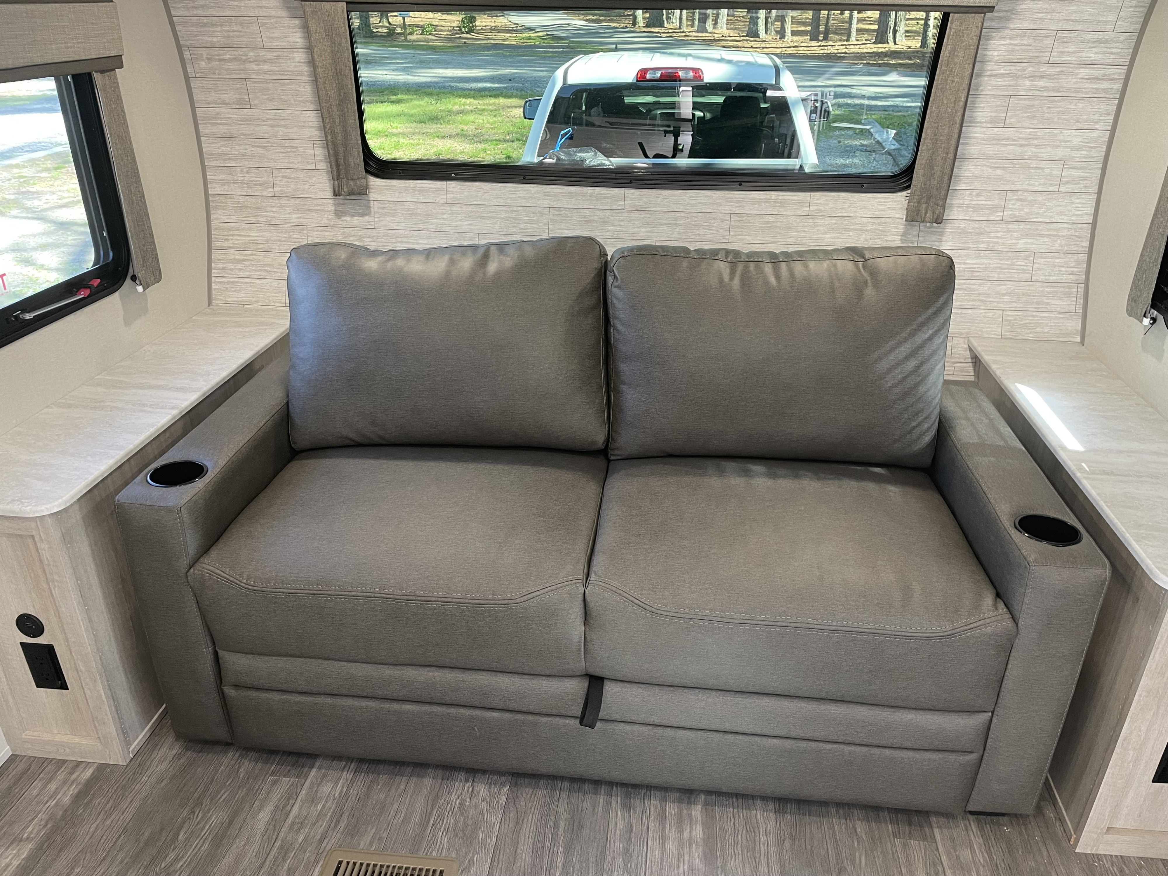 Comfortable grey sofa with cup holders inside an RV rental, offering a view of a truck and trees through large windows.. Forest River Aurora 2024